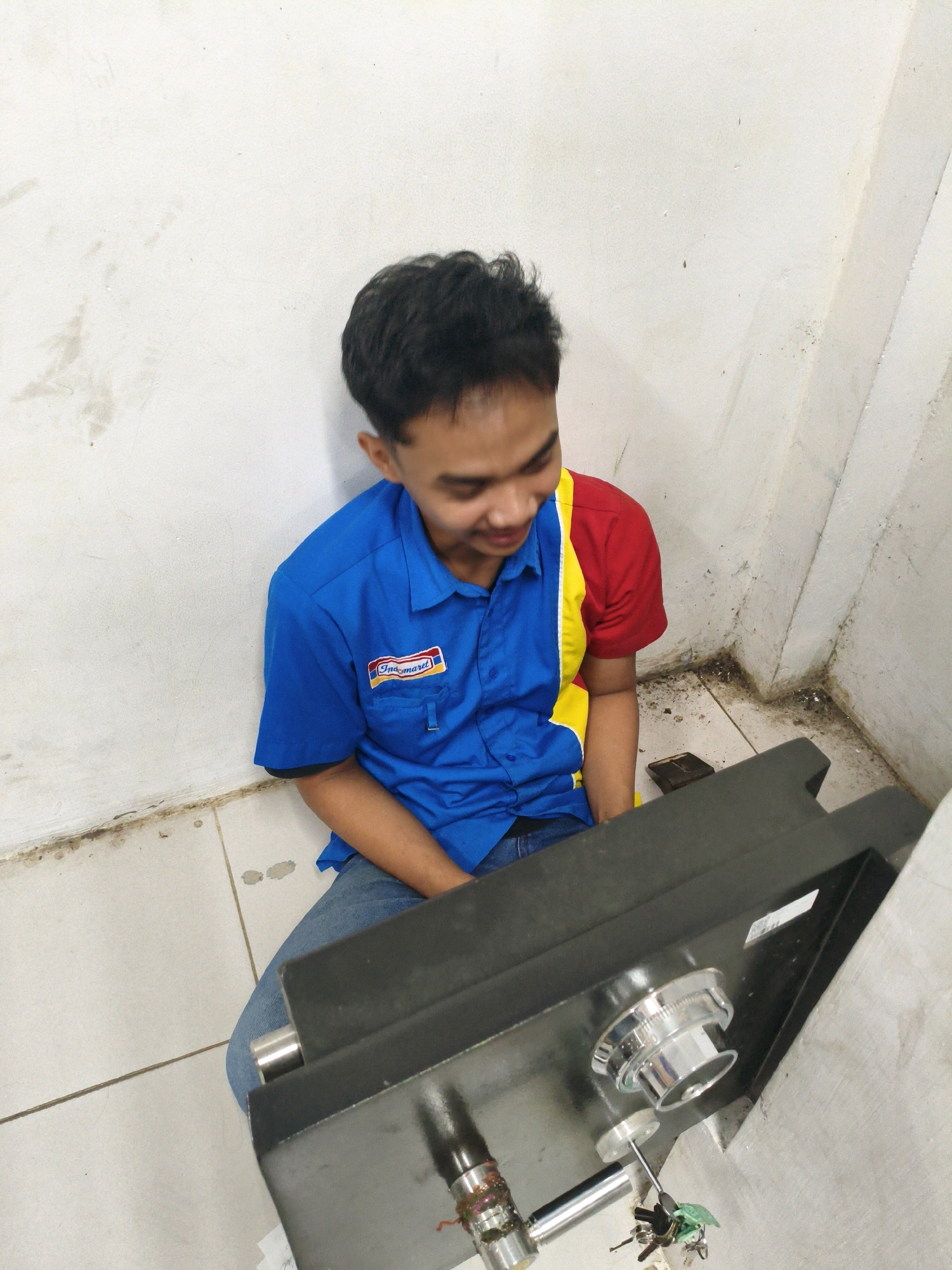 A young man wearing a blue shirt with red and yellow accents sits on the floor next to a dark metal safe. The shirt has a white and red logo on the left breast pocket. He is looking down and to his right, with his head tilted and a slight smile on his face. The safe has a silver dial and is partially open. A set of keys is attached to a small green item and is hanging near the safe's dial. The man is wearing blue jeans. The environment appears to be an interior space with white walls and a tiled floor. The walls show some signs of age and dirt. The scene is captured from a high angle, looking down at the man and the safe. The location is identified as Sumberejo Kulon, Indonesia.