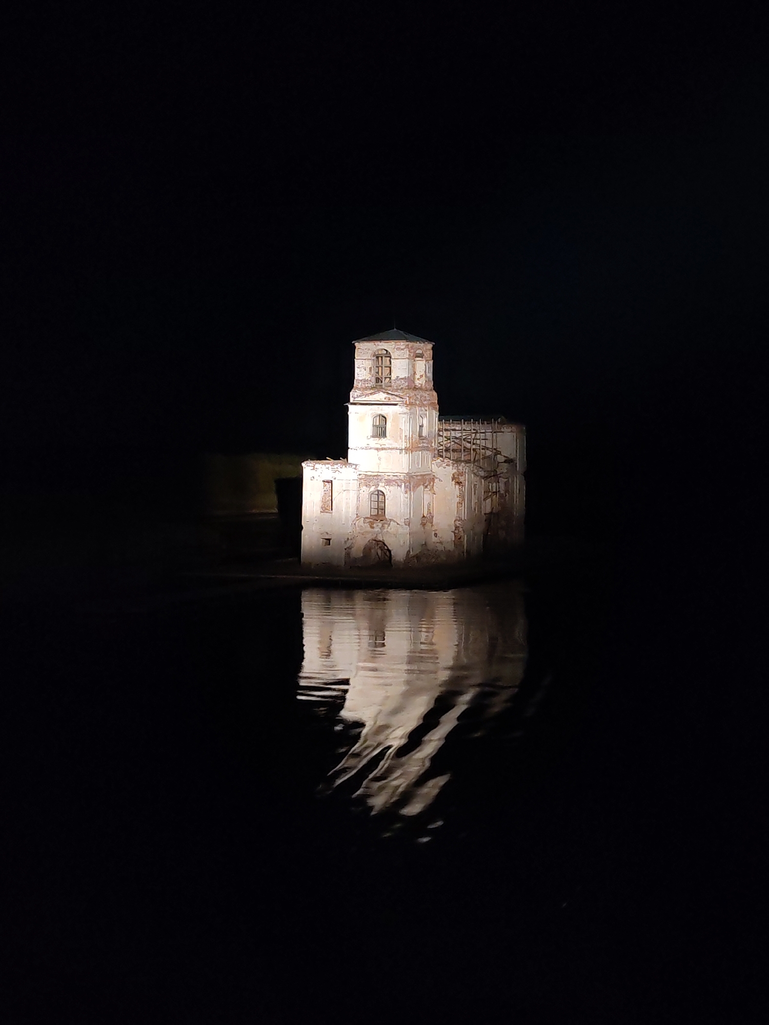 The image depicts the partially submerged, ruined Church of the Nativity in Krokhino, Russia, at night. The church is the only illuminated object in the scene, with its reflection visible in the surrounding water. The structure has a multi-tiered tower and noticeable damage to its walls and upper levels. Scaffolding is visible on one side. The rest of the image is dark and appears to be water.