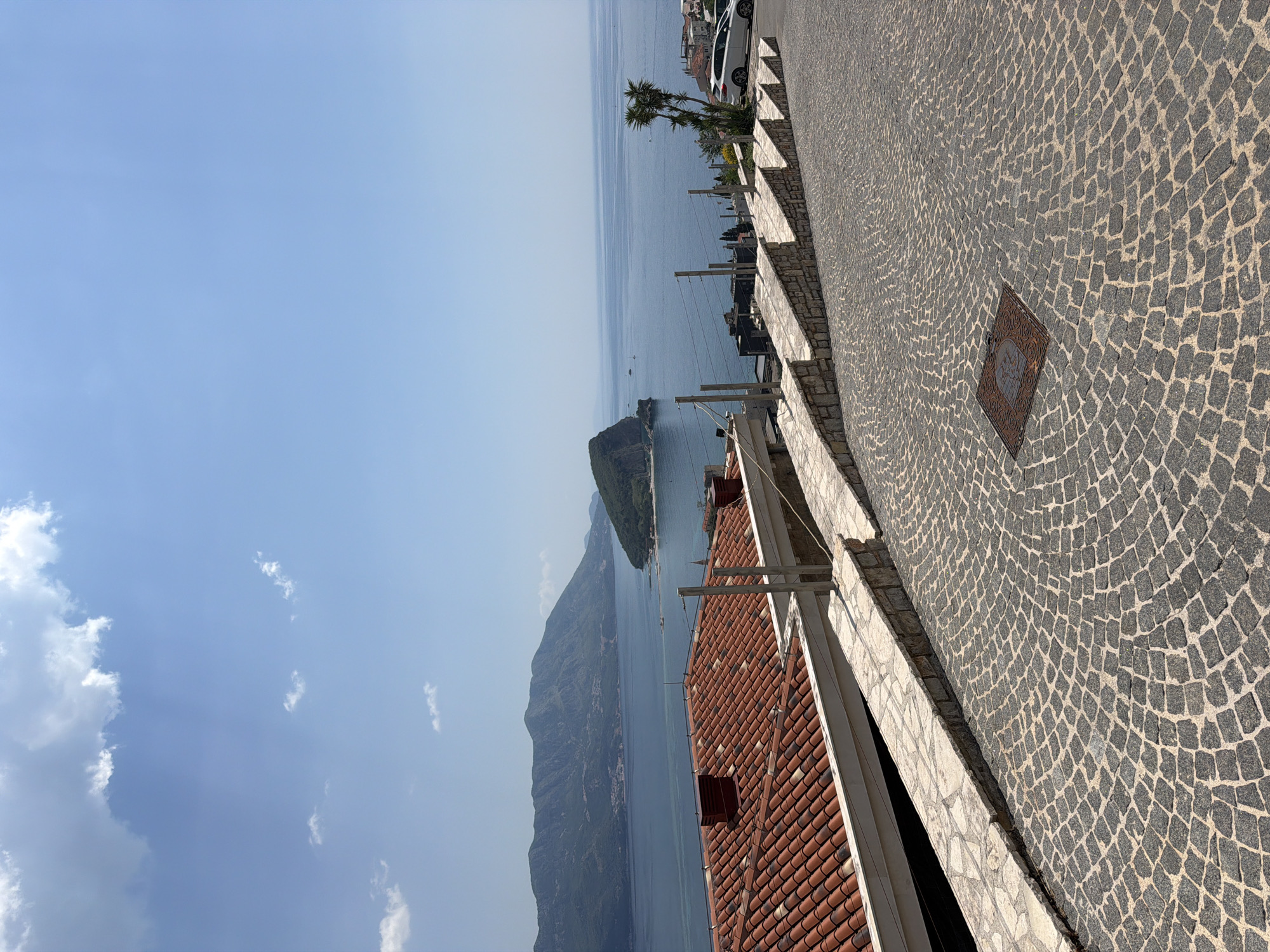 The image presents a view of Lake Garda, Italy from a high vantage point. The sky is predominantly blue with scattered clouds. A mountainous peninsula stretches into the lake, with a small island visible further out. In the foreground, there is a red-tiled roof. Adjacent to the roof is a paved area with a cobblestone pattern. A low wall separates the paved area from the roof. The photo is taken in a slightly tilted position. The location is not Budva, Montenegro; it is Lake Garda, Italy.
