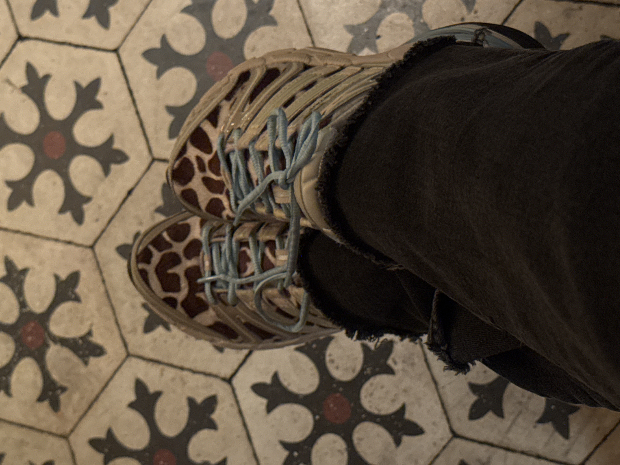 An overhead shot shows a person standing on a patterned tile floor. The person is wearing black jeans with frayed edges and a pair of tan and brown sneakers with light blue laces. The tiles are hexagonal, with a white background and a black and red floral pattern. The location is in Tel-Aviv, Israel.