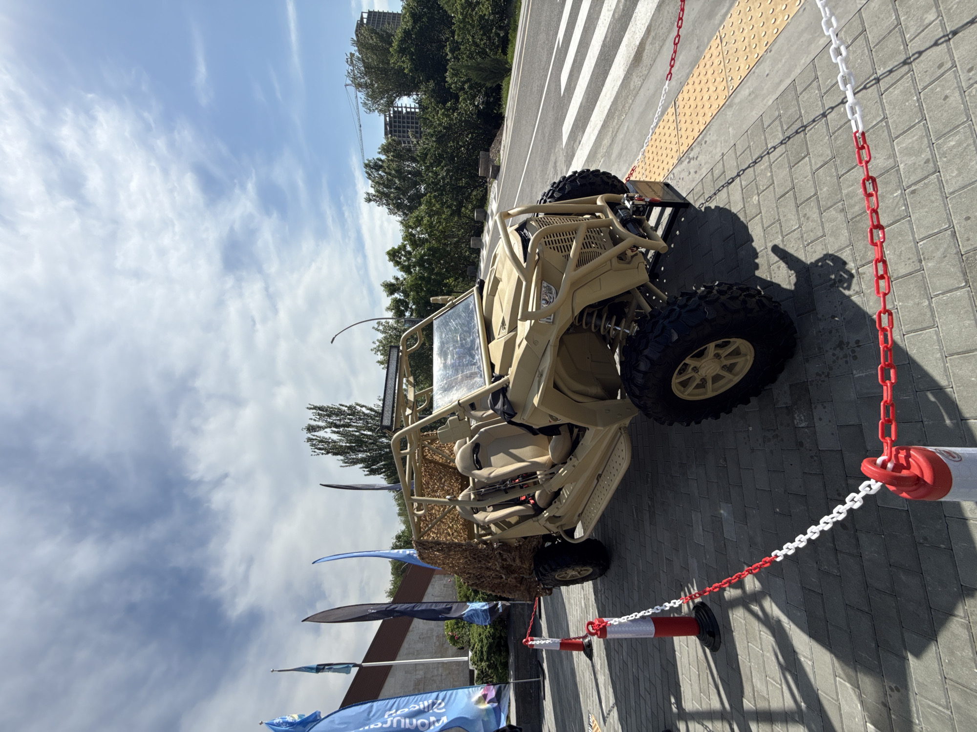 An outdoor scene features a tan-colored military or off-road utility vehicle, possibly a UTV or ATV, positioned on gray paver stones. The vehicle is equipped with large, knobby tires, an exposed roll cage, and two visible bucket seats. It is bordered by red and white plastic chains connected to matching bollards, suggesting it is on display or cordoned off.
In the background, a road with a white zebra crossing is visible, adjacent to a yellow tactile paving strip. Beyond the road, there are abundant green trees and a building, which appears to be under construction, with a crane visible. The sky is clear and blue with scattered white clouds, indicating bright, sunny weather, likely mid-day or early afternoon given the distinct shadows cast by the vehicle and chains.
To the left of the vehicle, several blue and black flag banners are displayed. Partial text is visible on one blue flag, including "MON..." and "...um," with "...RUS" below. Another banner shows fragments of words like "sile" and "sium." The setting appears to be an exhibition or event space. A shadow resembling a person holding an object, possibly a camera or phone, is cast on the pavement near the chains. The overall atmosphere is calm and orderly.