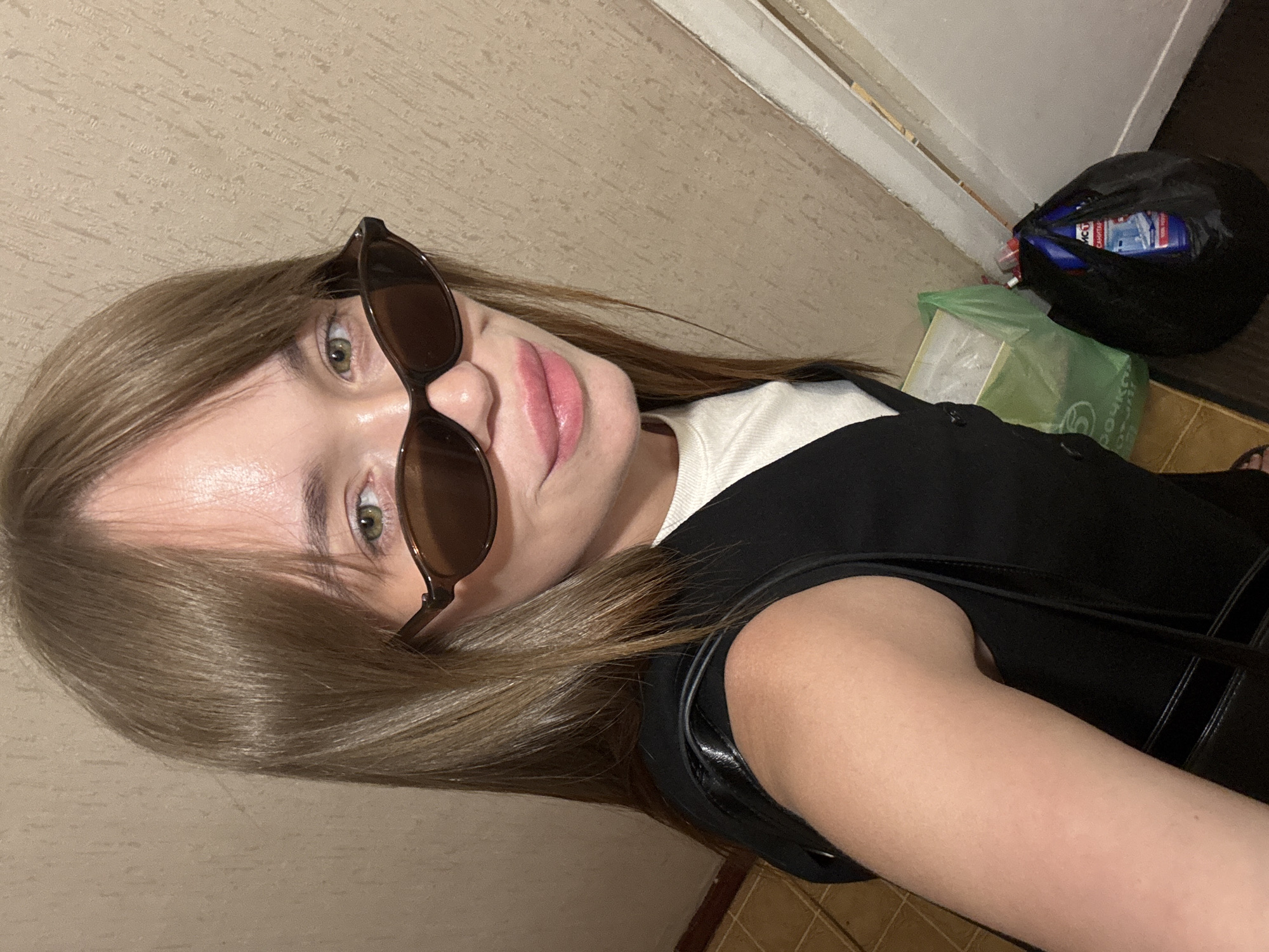 A woman with light brown hair and green eyes is posing for a selfie. She is wearing sunglasses and a black top over a white shirt. In the background, there is a doorway and a black garbage bag with a blue bottle visible inside. A green bag is also visible near the garbage bag. The location appears to be indoors, with beige walls and flooring. No definitive details pinpoint the location to Chaikovsky, Russia.