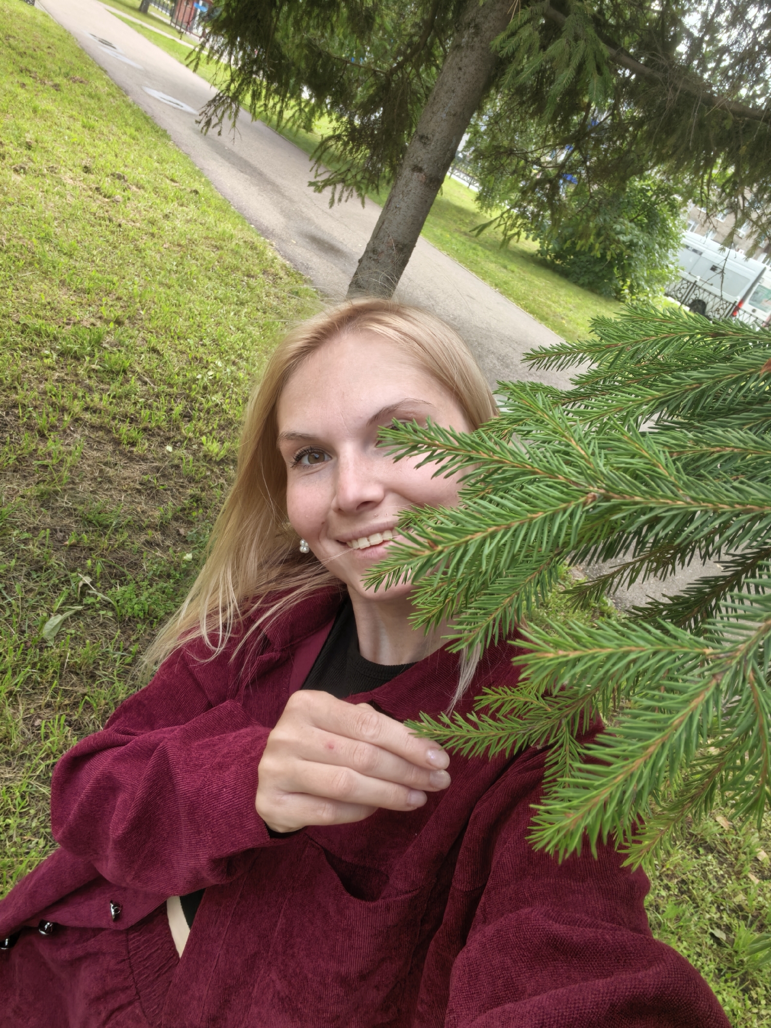 A woman with blonde hair and fair skin is pictured from the chest up, partially obscured by a pine branch. She is wearing a burgundy corduroy jacket over a black shirt. The woman is smiling, with her head tilted slightly to the right. Her left hand is resting on the pine branch, and she is looking towards the camera. The background is a grassy area with a paved path and trees. The location is identified as Sterlitamak, Russia.