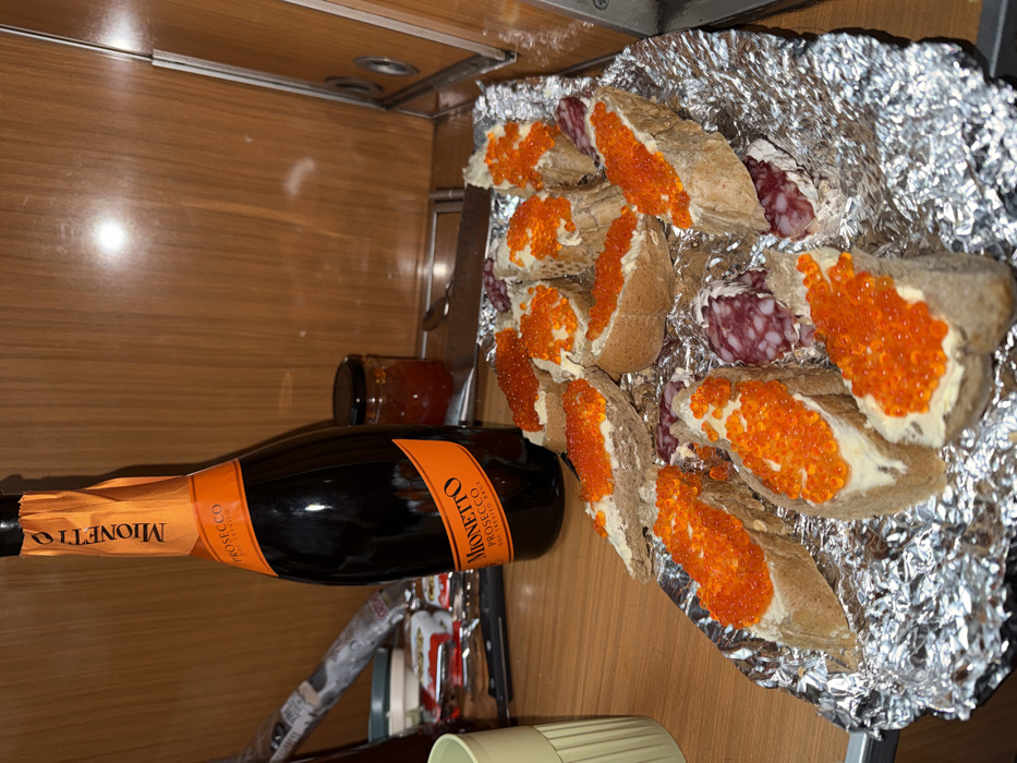 A table displays a bottle of Mionetto Prosecco, a small jar of jam, and a tray of canapés. The canapés feature sliced baguette bread topped with butter and orange caviar. Some slices of salami are mixed in with the bread. The presentation is on a sheet of crumpled aluminum foil. The table has a wood-grain finish.