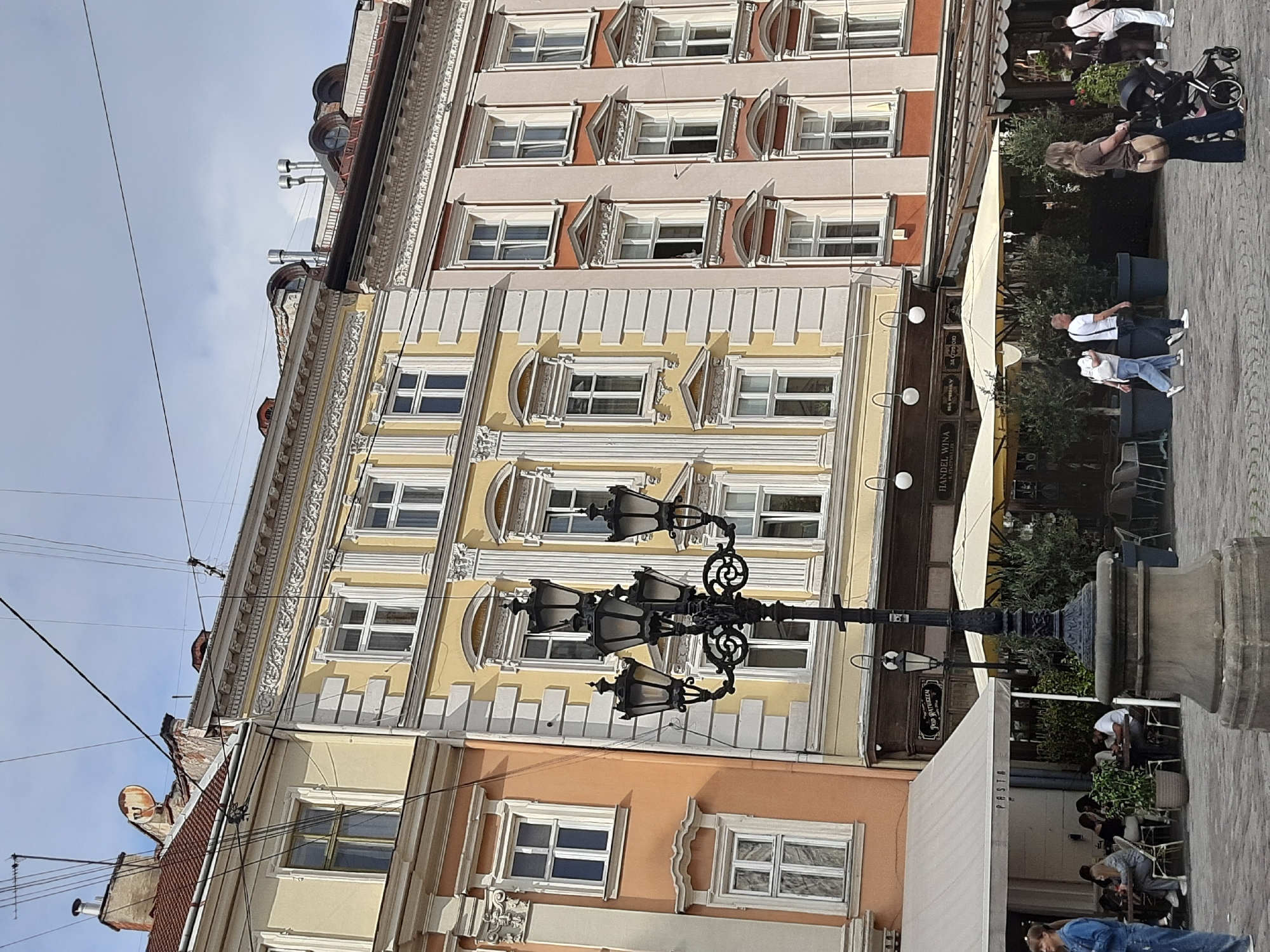 The image shows a street scene in Lviv, Ukraine, on a cloudy day. In the foreground, there is a cobblestone street with a large, ornate lamppost. To the left, an outdoor seating area of a restaurant named "PASTA" is visible, with several people seated at tables. Beyond the restaurant, there are awnings over outdoor seating for other establishments. In the background, there are multi-story buildings with yellow and pink facades, featuring numerous windows and decorative architectural elements. People are walking on the street, including a woman with a stroller. The overall atmosphere is that of a bustling European city square.