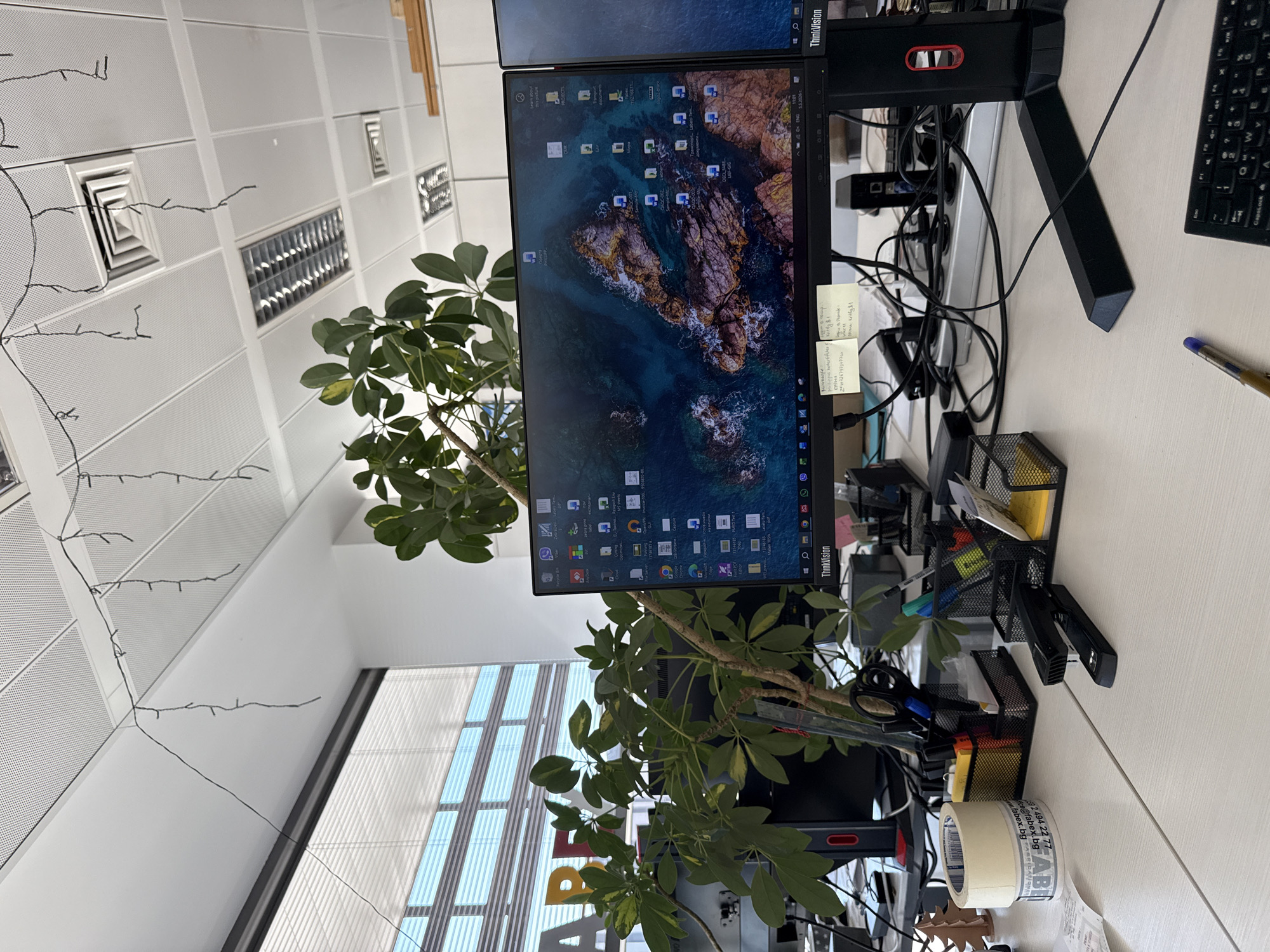 Here is a matter-of-fact description of the scene:
The image shows a desk in an office setting, likely in Sofia, Bulgaria, judging from the tape with a Bulgarian domain on it. A computer monitor stack is the central focus, displaying a nature-themed background, with sticky notes attached to the side. A plant stands next to the monitors. A keyboard and a pen are on the desk. A wire management system is in place to keep the wires organized. The desk also contains desk organizers holding pens and office supplies such as scissors and paper clips. The office has drop ceilings with recessed lighting. A window with blinds is located in the background. The image is rotated for an unclear reason.
