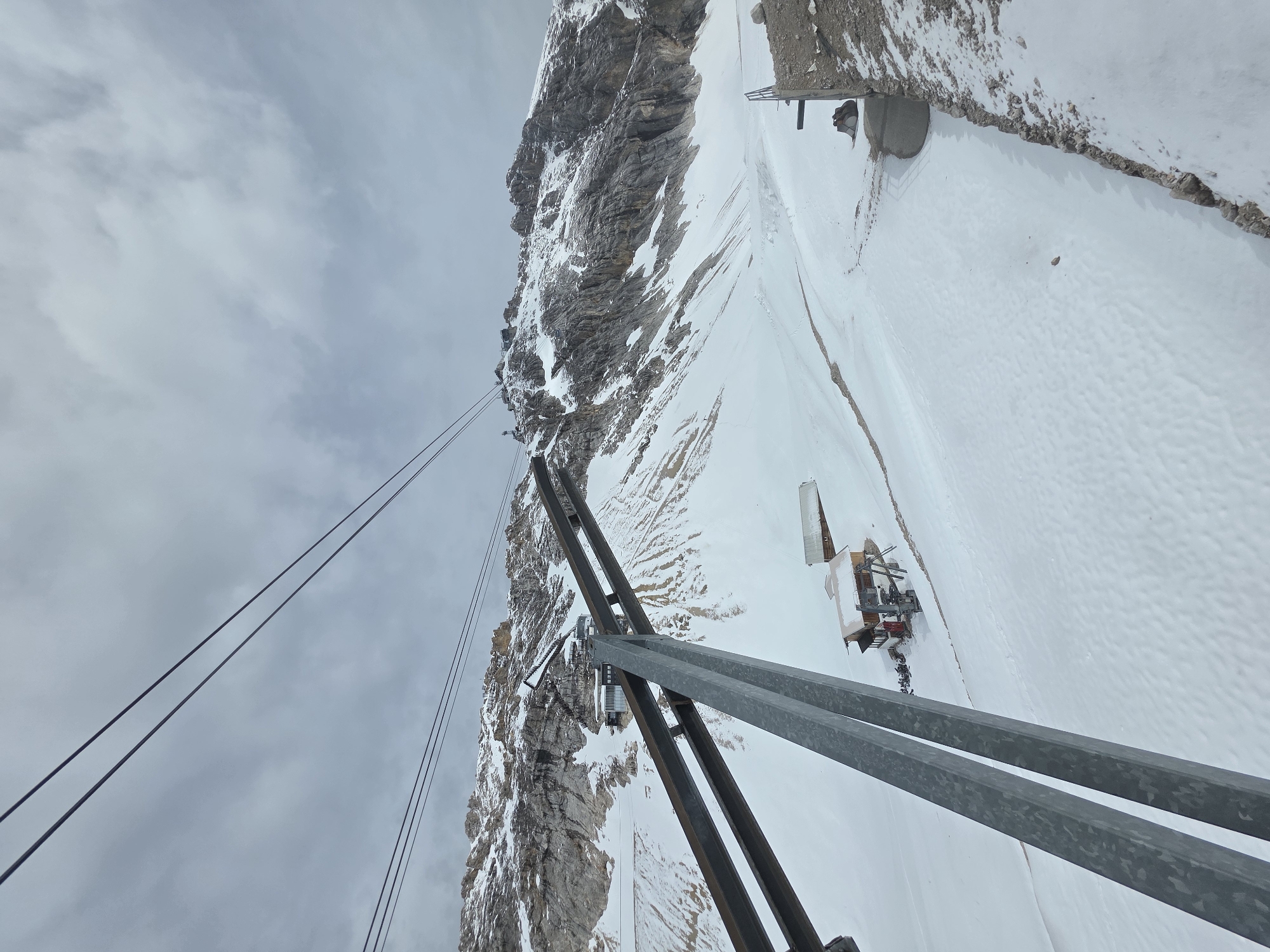 The image presents a high-angle, downward-looking view of a snow-covered mountainous landscape from what appears to be a cable car or gondola system, located in the city of Unknown, Unknown. In the immediate foreground, a robust metallic support structure, likely part of a cable car pylon, extends diagonally from the lower left towards the upper center, partially obscuring the view. Four distinct dark cables stretch across the upper portion of the frame, disappearing into the overcast, grey sky which exhibits varying densities of cloud cover.
The midground is dominated by an expansive snowfield, displaying subtle undulations and faint track marks, indicating human activity. Nestled on this snow-covered slope are several structures. Towards the lower right, a small, rustic wooden building with a brown roof and red accents is visible, accompanied by some machinery or equipment outside. Further up the slope and to the left, a more contemporary multi-level building with a dark roof and prominent glass sections is integrated into the terrain, connected by a series of stairs ascending to an unseen point higher on the mountain. Another smaller building is partially visible behind the rustic cabin.
The background consists of rugged, rocky mountains, heavily dusted with snow and ice, revealing a complex geology of dark rock faces interspersed with white snow patches. A mountain peak towards the upper left exhibits additional infrastructure, including what appear to be multiple pylons or small structures, suggesting a summit station or observation point. The overall environment is one of a high-altitude, cold, and predominantly natural setting, modified by infrastructure for mountain access and tourism. No human figures are discernible in the scene.