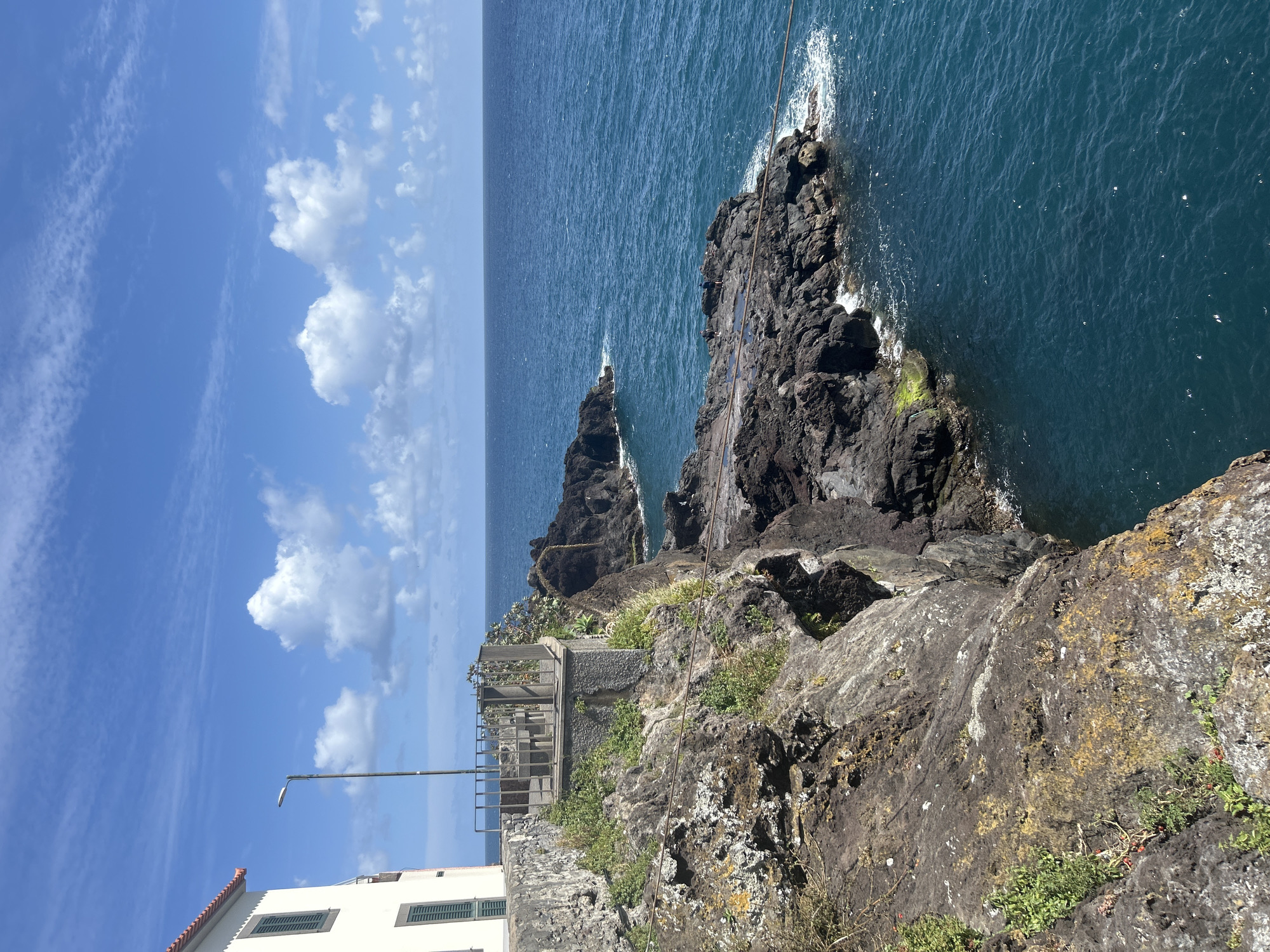 The image presents a vertical shot of the coastal area in Câmara de Lobos, Portugal. The scene captures a rocky cliff face meeting the dark blue ocean, with two prominent rock formations jutting out of the water. The cliff face is textured with varying shades of gray and black rock, interspersed with patches of green vegetation and light-colored lichen. A metal railing structure is visible on a constructed stone platform, situated on the cliff's edge, indicating a viewpoint or walkway. Below the platform, a white building with dark green window shutters and a red tiled roof is partly visible. A street lamp rises from the building's side. The sky is blue with scattered white cumulus clouds. The ocean extends into the horizon, marked by gentle ripples and the interplay of light. A cable-like structure runs down the side of the rock formation.