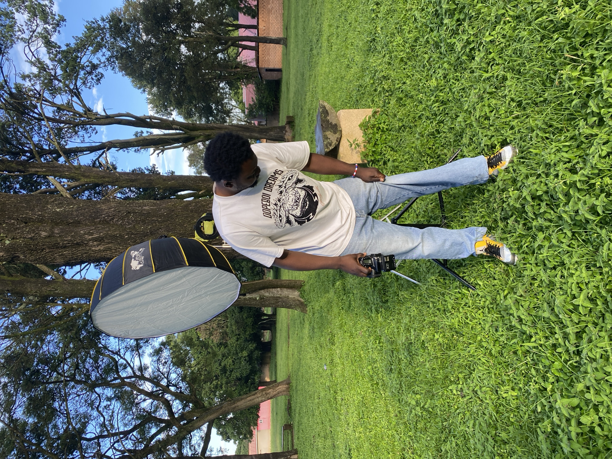 A young Black man with dark curly hair is standing in a grassy park in Jos, Nigeria. He is wearing a white t-shirt with a graphic print, blue jeans, and yellow and black sneakers. He is holding a camera in his right hand and looking down at his jeans. To his left, a large, black and gray photography reflector is propped up on a stand. In the background, there are several large trees, a red-roofed building, and a brick structure. The sky is blue with scattered white clouds. The overall scene suggests a casual outdoor photography session.