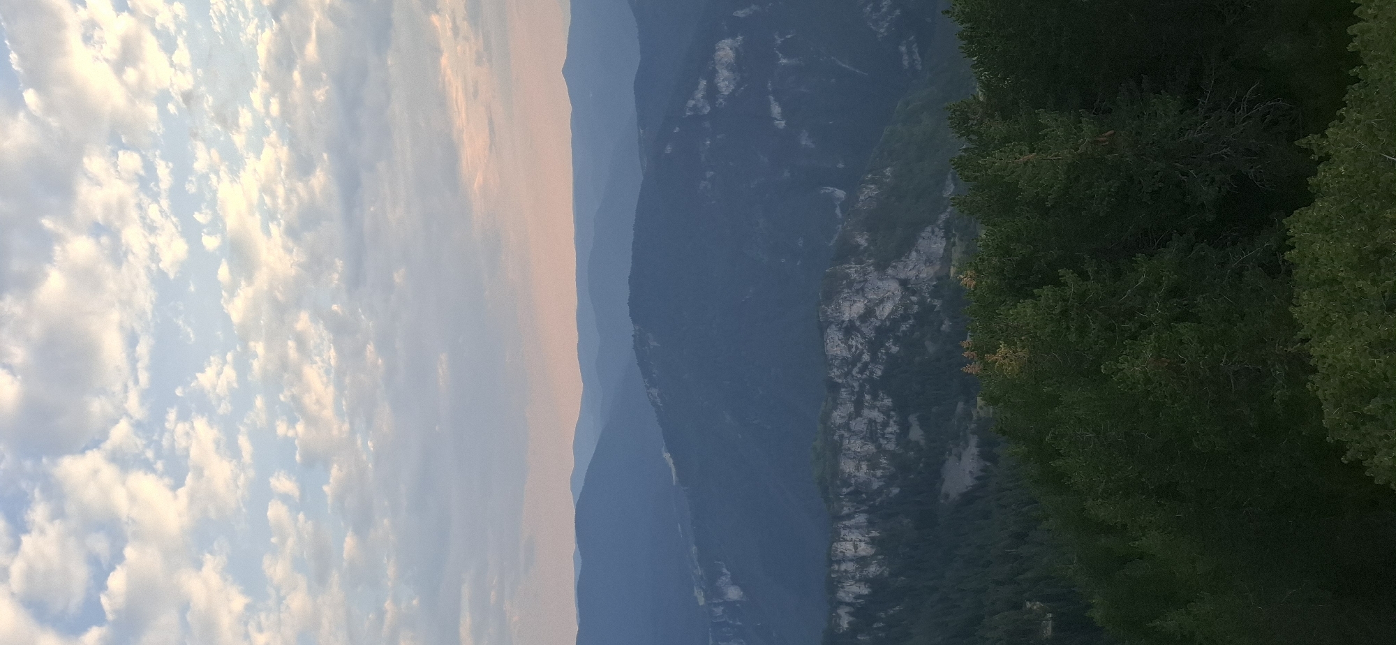 This is a vertical, medium-shot, eye-level photograph taken from a high vantage point overlooking a mountainous landscape at dusk. The sky occupies the top third of the image and is filled with scattered, wispy clouds, tinged with soft pinks and oranges from the setting sun, contrasting with the pale blue sky peeking through. Below the sky, a series of receding mountain ranges are visible, each fading into a deeper shade of blue and purple with distance. The closest mountain range in the foreground is densely covered with dark green coniferous trees. Prominent on the mid-ground of the image is a dramatic, rugged cliff face with a stark, rocky texture and lighter colored stone, dotted with patches of green vegetation. The overall impression is one of natural grandeur and tranquility. The image does not depict any people or man-made structures. It is a landscape photograph of the mountains near Gyovren, Bulgaria.