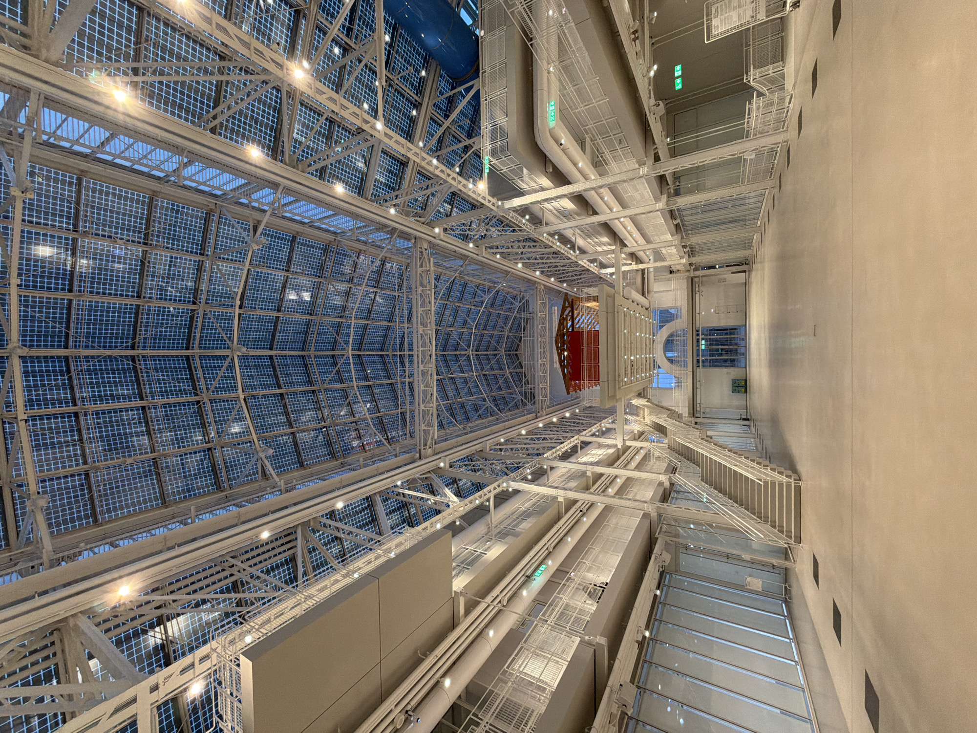 The image depicts an interior space, likely a large atrium or hall, with a complex ceiling structure. The ceiling is primarily made of a grid-like pattern, possibly glass panels with metal framing. There are numerous lights installed within the ceiling, illuminating the space. Below the ceiling, there's a network of pipes, ducts, and walkways, suggesting a modern industrial or institutional building. The walls are plain and off-white, with stairs and landings visible. The overall impression is of a spacious, well-lit, and modern architectural design. The location appears to be indoors. The city and state are unknown.