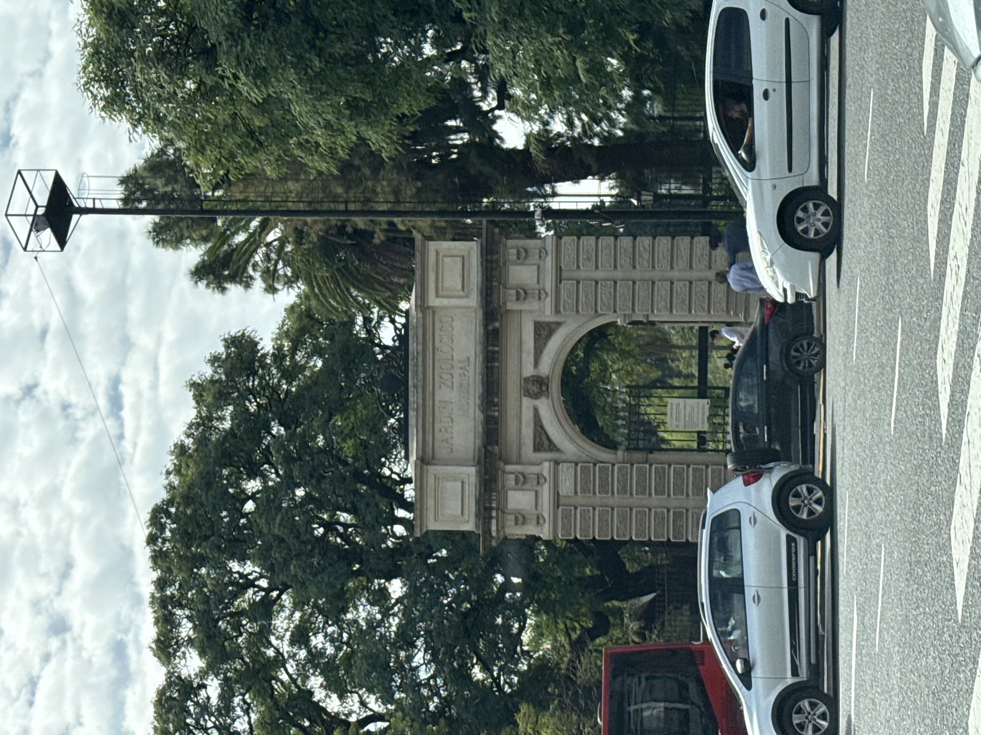 This image shows the entrance arch of the Buenos Aires Zoo, now known as Ecoparque Interactivo, in the Palermo neighborhood of Buenos Aires, Argentina. The archway is a beige, ornate structure. A tall lamppost with a cubic fixture stands to the left of the arch. In the foreground, several cars are visible on a city street with pedestrian crosswalks. The image includes trees, and the sky is visible with scattered clouds. People can be seen in and around the cars and the zoo entrance.