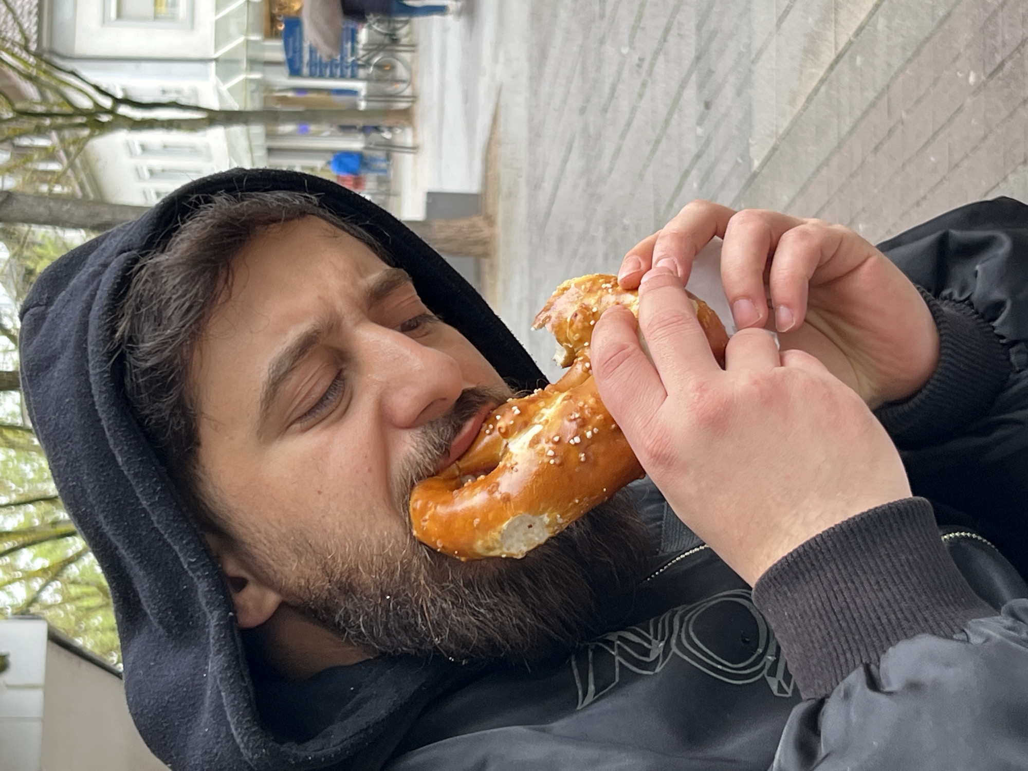 A bearded man in a black hooded jacket is shown eating a pretzel outdoors. He is holding the pretzel with both hands, about to take a bite. The pretzel has salt on it. He is wearing a black jacket with a ribbed cuff. The background appears to be a street scene in the city of Kehl, Germany, with buildings and trees visible. The image is rotated counter-clockwise.