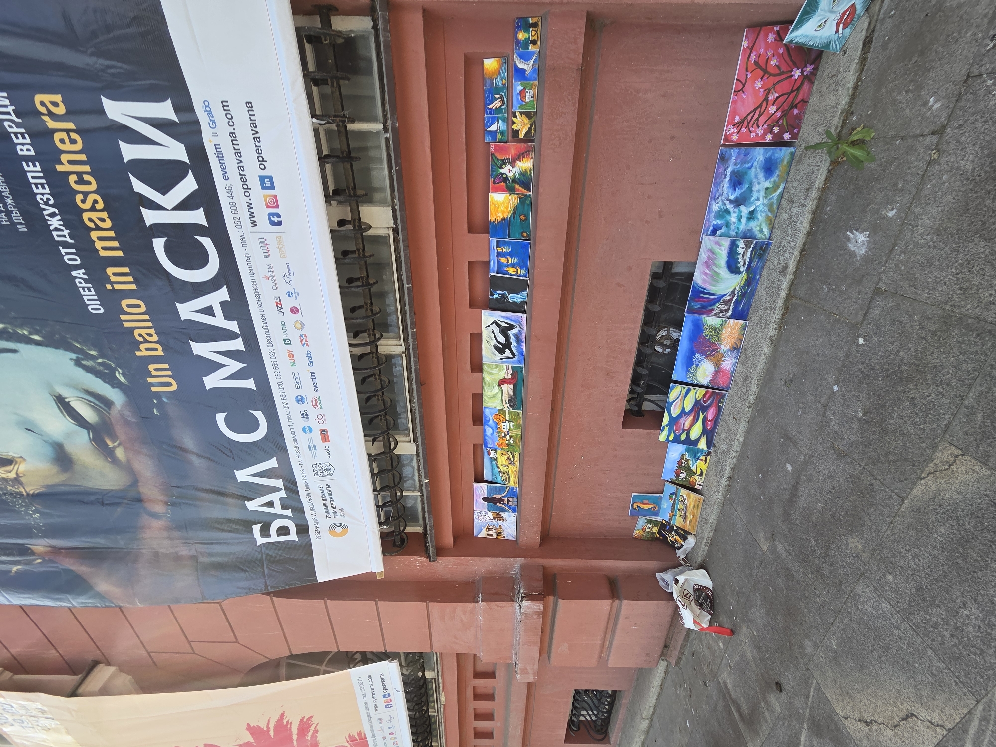 The image captures an outdoor scene in Varna, Bulgaria, featuring a large promotional banner alongside an informal display of numerous small paintings.
The most prominent feature is a large, vertically oriented banner advertising "Un ballo in maschera" (A Masked Ball) by Giuseppe Verdi, translated to "БАЛ С МАСКИ" in Bulgarian. The banner, predominantly black and white with golden and blue accents, shows a partial image of a person in a mask or ornate costume. Visible text on the banner includes contact information for ticket reservations and sales: "ОПЕРА ОТ ДЖУЗЕПЕ ВЕРДИ", "Un ballo in maschera", "БАЛ С МАСКИ", phone numbers like "052 665 020", "052 665 022", and "052 608 446", and the website "www.operavarna.com" along with social media handles for "operavarna" on Facebook, Instagram, and LinkedIn. It also lists various sponsors and partners such as Eventim, Grabo, NJOY RADIO, Jazz FM, Classic FM, and Magic.
Below and to the right of the banner, an array of small, colorful paintings are displayed along a reddish-brown building facade and on the adjacent gray sidewalk. Some paintings are neatly arranged on narrow ledges or recessed areas of the wall, while others are simply laid out on the ground. The subjects of these artworks are diverse, including vibrant underwater scenes with coral and fish, seascapes with boats and lighthouses, animals (a cat, a dolphin, a seahorse), floral patterns like cherry blossoms, abstract designs, and figurative works. The informal arrangement suggests an artist or vendor displaying their work for sale.
The building itself is a multi-story structure with a distinctive reddish-brown facade, possibly stucco or brick, featuring some architectural detailing and window grates. The ground beneath is a gray concrete sidewalk. A discarded white plastic bag with some items inside is visible near the paintings on the sidewalk, and a small green plant sprouts from a crack in the pavement, adding a touch of natural detail to the urban setting. The scene appears to be taking place during daylight hours, under clear weather conditions.