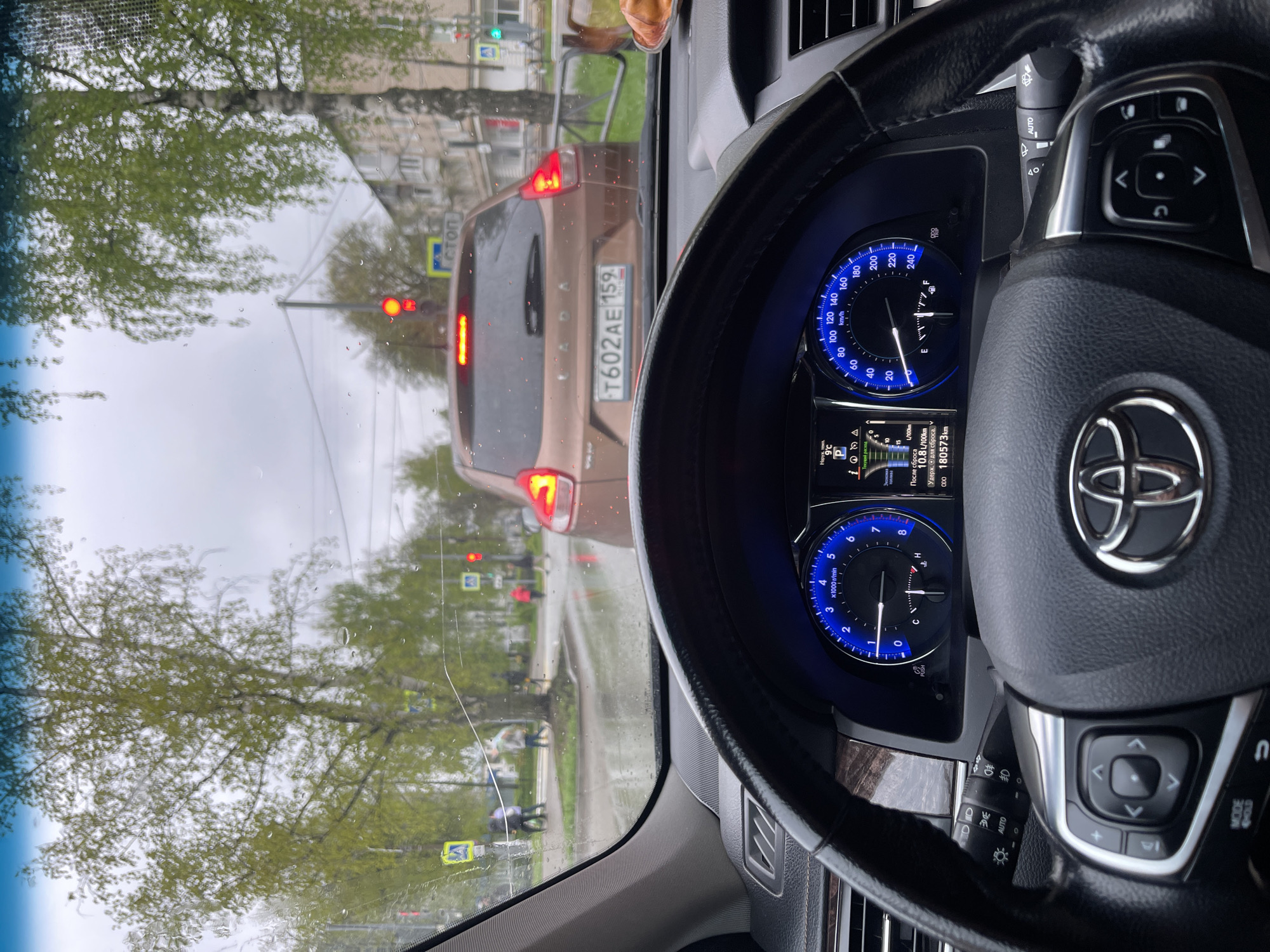 The image, taken from inside a car in Chaikovsky, Russia, shows the driver's perspective of a street scene. The dashboard and steering wheel of a Toyota are prominently featured in the foreground. Through the windshield, a brown Lada car with the license plate "T602AE 159" is visible, stopped at a traffic light. The traffic light is red. Pedestrian crossing signs are visible on the side of the road. Trees line the street, with their spring foliage visible. The weather appears overcast. The car's dashboard displays information including the outside temperature ("9°C"), fuel consumption ("10.8L/100km"), and odometer reading ("180573km").