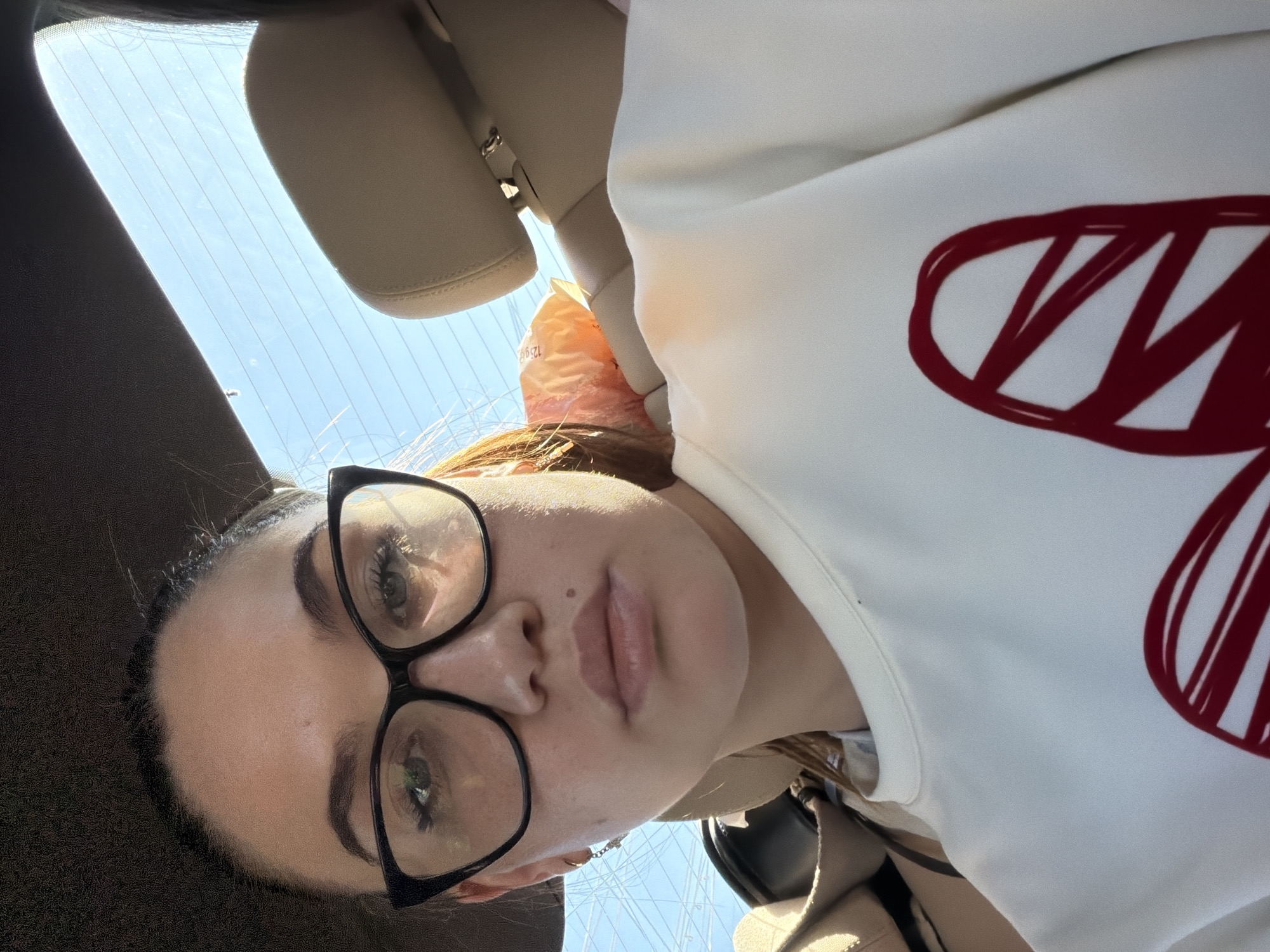 A woman with dark hair pulled back and wearing black-rimmed glasses is looking directly at the camera. She has clear skin with a small mole on her right cheek. Her lips are slightly parted, and she is wearing a white sweatshirt with a large red heart design. She is seated in a car, with a light-colored headrest visible behind her and a bright blue sky with faint horizontal lines through a car window above. A bag of orange snack chips is visible on the passenger seat next to her. The interior of the car appears to be a neutral beige color. The overall lighting suggests daytime. There is no specific indication of the location in the city of Zorleni, Romania.