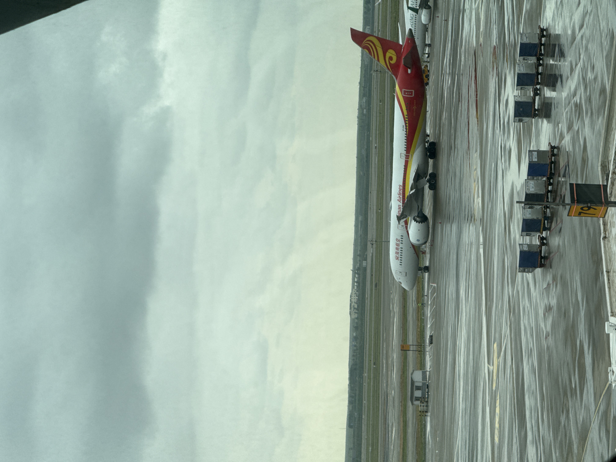 A wide shot captures a Hainan Airlines Boeing 787 Dreamliner airplane on a wet tarmac at Pudong International Airport in Shanghai, China. The sky is overcast and cloudy, and the tarmac is wet, reflecting the gray light. The plane is the central subject, with its distinctive red and gold livery visible. The phrase "Hainan Airlines" is written in English on the fuselage, and Chinese characters are also present. The words "DREAMLINER" are printed near the rear of the aircraft. In the foreground, several baggage carts with blue and gray containers are parked on the wet tarmac. In the background, other planes and airport buildings are visible, along with a distant line of trees and structures. The overall impression is of a typical airport scene on a cloudy day, with the focus on the large aircraft.