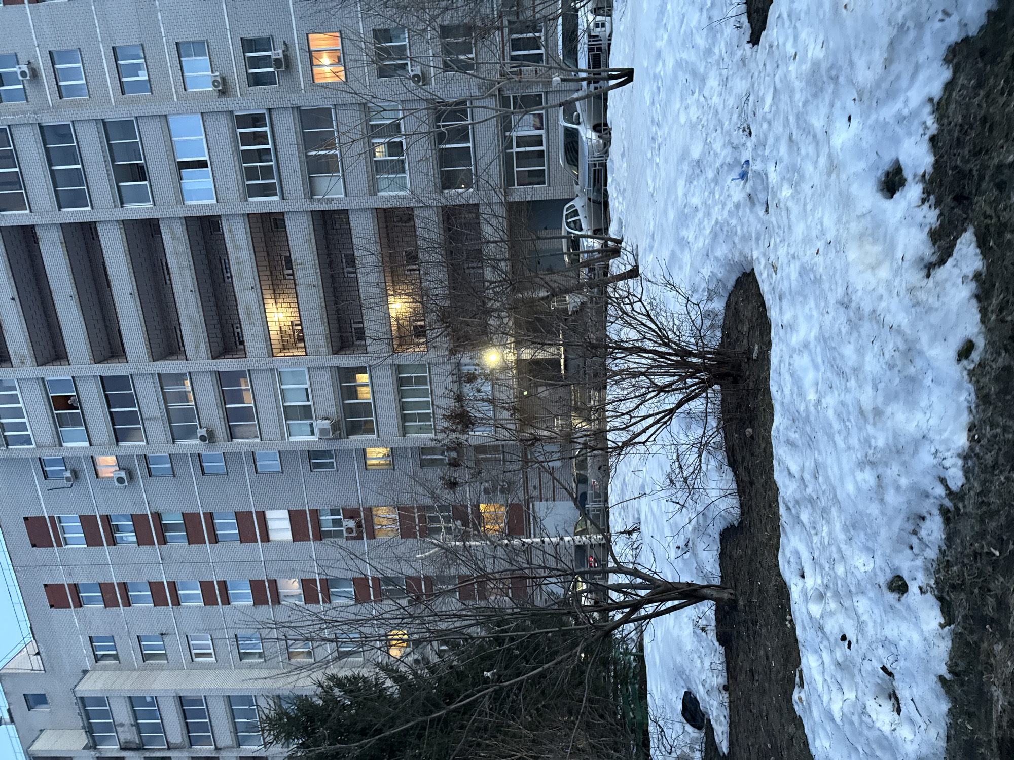 The image shows a multi-story gray brick apartment building in Izhevsk, Russia. The building has rows of rectangular windows, some with lights on inside. There are bare trees in front of the building, and patches of snow on the ground. Several parked cars are visible in front of the building. The scene appears to be in early spring, with melting snow. The sky is overcast.