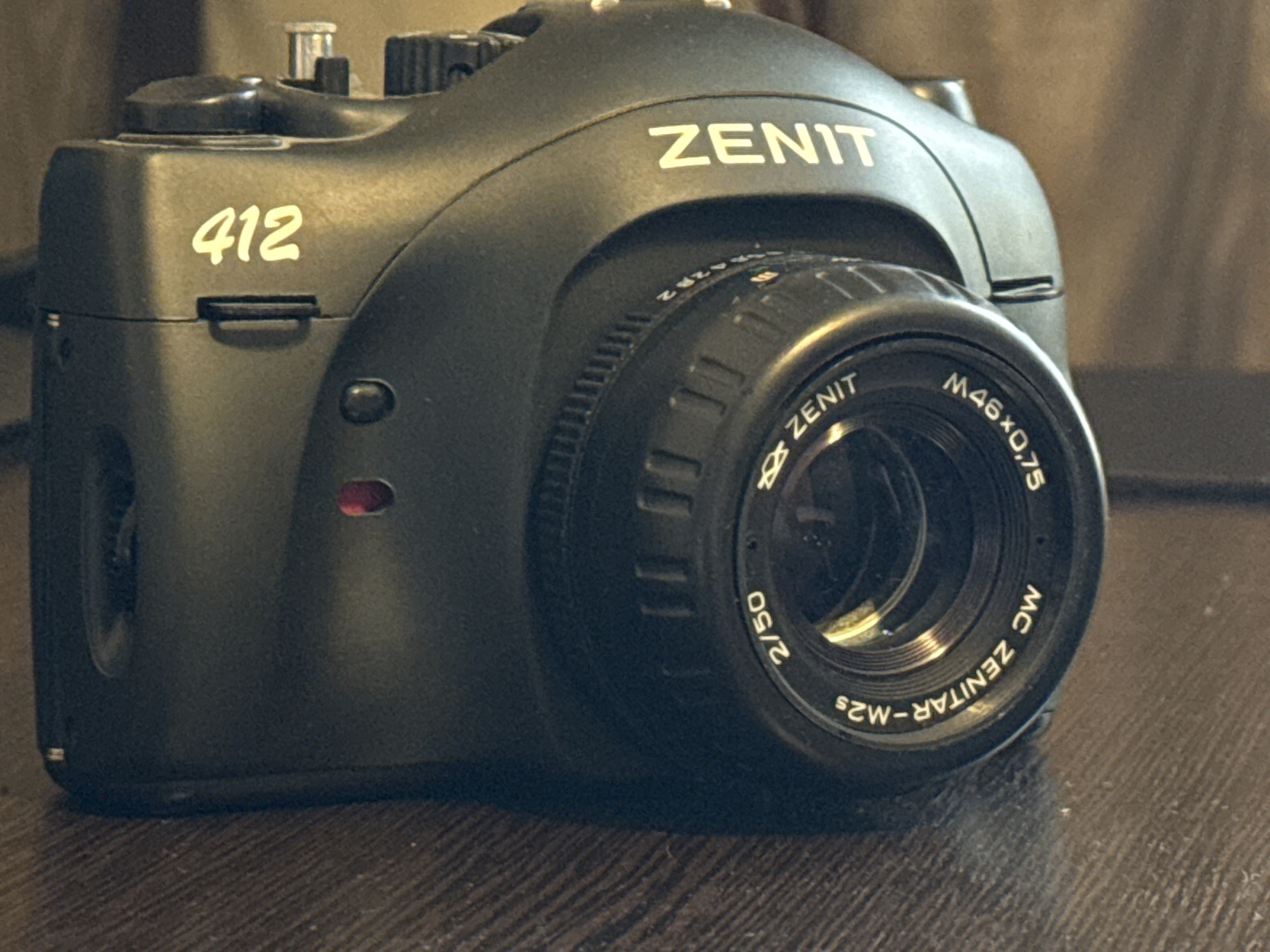 The image shows a close-up, slightly angled view of a black Zenit 412 camera with a lens attached. The camera is placed on a dark wooden surface, likely a table. The background is a blurred, textured brown material, possibly a curtain or fabric wall covering.
The camera body is predominantly black with some silver accents on the top. The model number "412" is visible in a lighter color on the upper left side of the camera body. The word "ZENIT" is prominently displayed in silver lettering on the top curve of the camera body, above the lens.
The lens attached to the camera is also black and features various inscriptions. Inscribed on the lens barrel are the words "ZENIT" and "M46x0.75". Below these, in a circular arrangement, is text that appears to read "MC ZENITAR-M2s" and "2/50". The aperture blades of the lens are partially visible and are a reflective metallic color.
There are no people or actions depicted in the image. The environment appears to be indoors, with artificial lighting contributing to the overall tone and shadows. The scene is static and focused solely on the camera. There is no indication of the city of Khimki, Russia, or any other specific geographical location.
