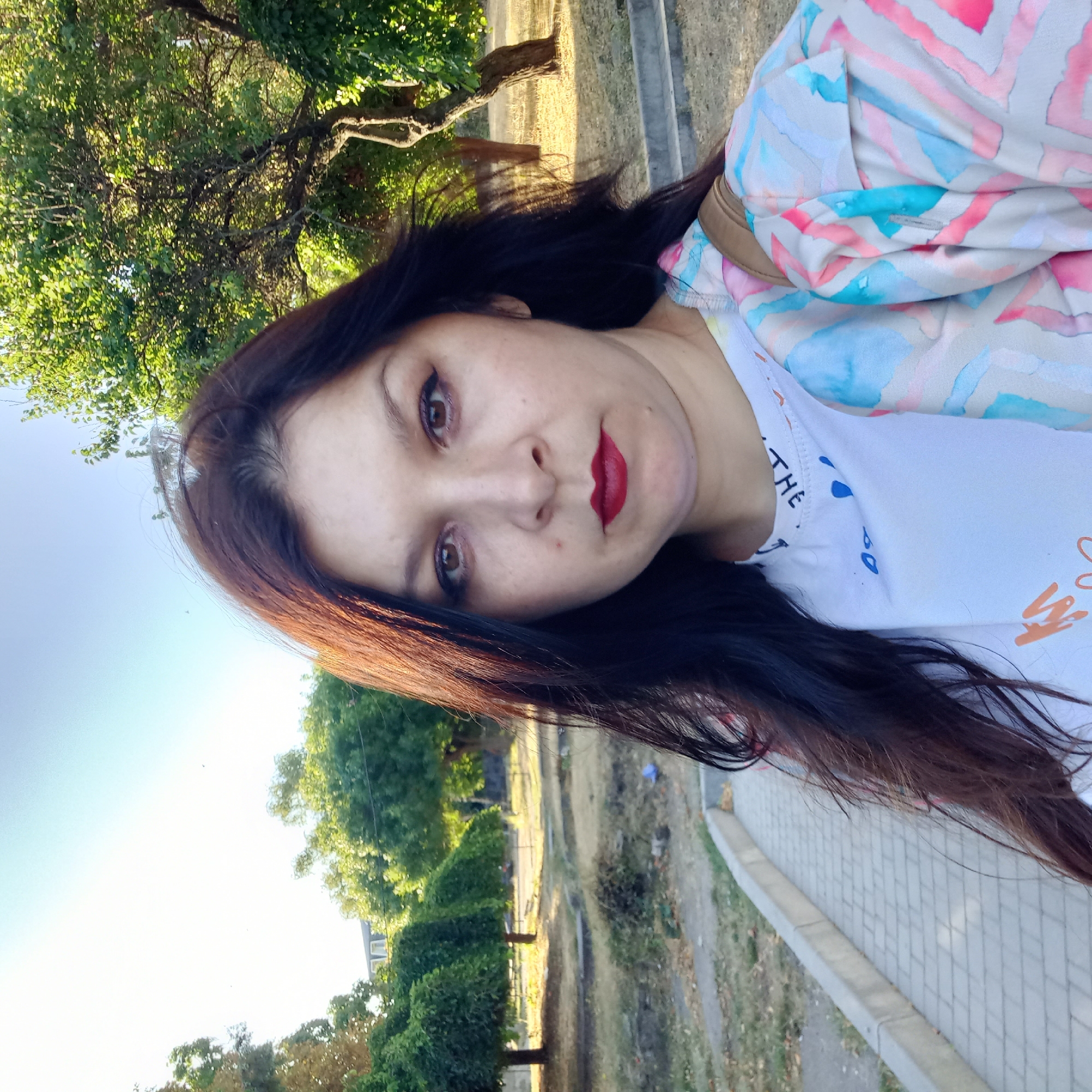 In an outdoor setting with a clear sky and ample daylight, a woman of East Slavic appearance stands and takes a selfie. She has long, dark brown hair, brown eyes, and fair skin, and is wearing dark eyeliner and red lipstick. She is dressed in a white t-shirt with a colorful, abstract watercolor pattern on her outer garment, which appears to be a light jacket or cardigan. She is also wearing a tan strap over her shoulder. The background shows a park-like environment with manicured trees, grassy areas, a paved walkway, and a glimpse of a building in the distance. The image may have been taken in Kropyvnytskyi, Ukraine, given that the subject is from that region. The overall mood of the image is serene and casual, with the subject appearing relaxed and confident.