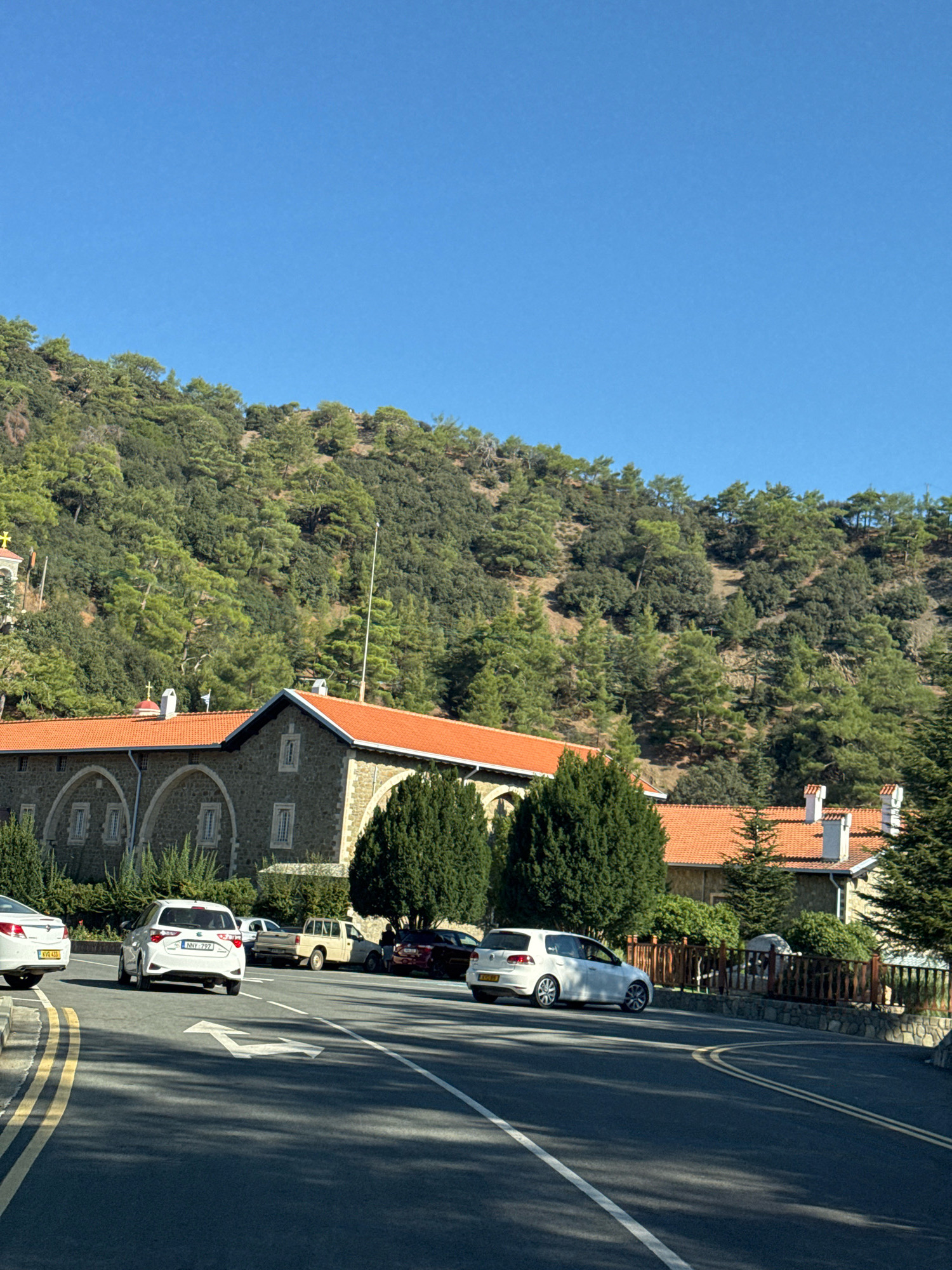 A street scene in Tsakistra, Cyprus, features a stone building with an orange tiled roof, possibly a monastery, nestled against a hillside covered in dense green trees. Several cars are visible on the asphalt road. In the foreground, a white Volkswagen Golf is parked on the right side of the road, and a white Suzuki is driving away from the camera in the left lane. Further down the road, other vehicles, including a pickup truck, are parked. The sky is a clear, bright blue.