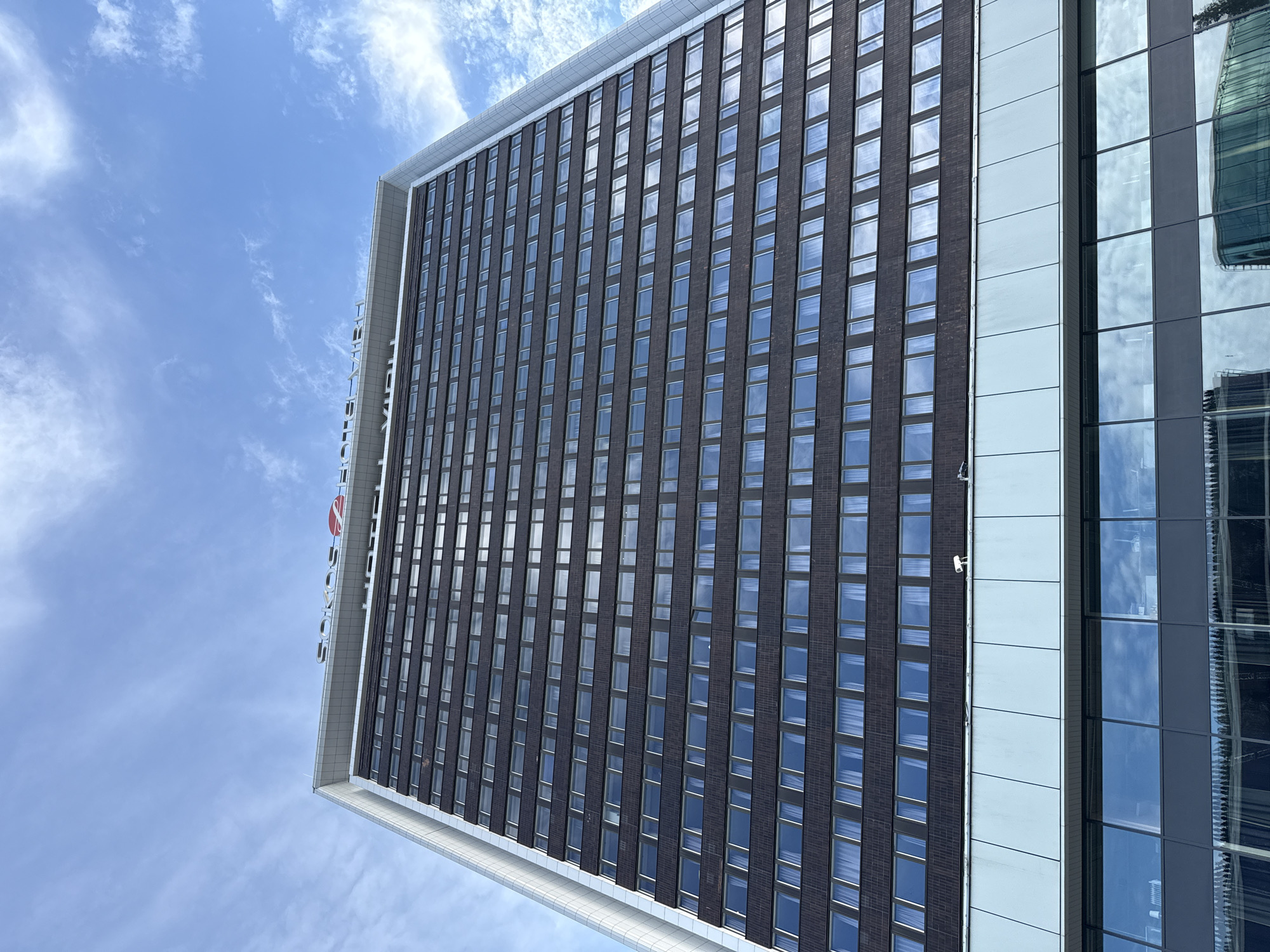 A low-angle, full shot captures a modern, dark-brown brick skyscraper against a bright blue sky with wispy white clouds. The building's facade is composed of numerous rows of rectangular windows, each with white curtains visible inside. The top of the building features a white cornice with the red and white "SOKOS" logo and a red heart symbol, along with the word "HOTEL VIRU" in silver lettering. A silver-colored vertical strip runs along the left edge of the skyscraper. Below the main body of the building, there is a lighter-colored section with a reflective surface, possibly glass, that mirrors the sky and some surrounding structures. The scene appears to be in an urban setting.