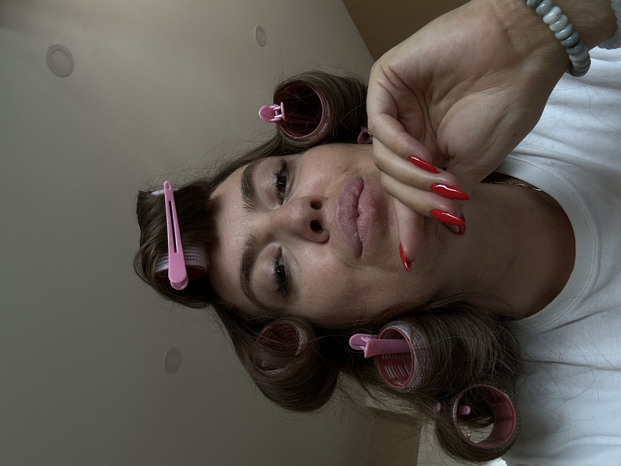 In an indoor setting, a woman with her hair set in pink rollers is looking at the camera. She is wearing a white t-shirt and has bright red manicured nails. Her hand is resting on her chin, and she appears to be posing for the photo. The background is a neutral-colored wall and ceiling with some recessed lighting. There is no visible indication of the location being in Batumi, Georgia.