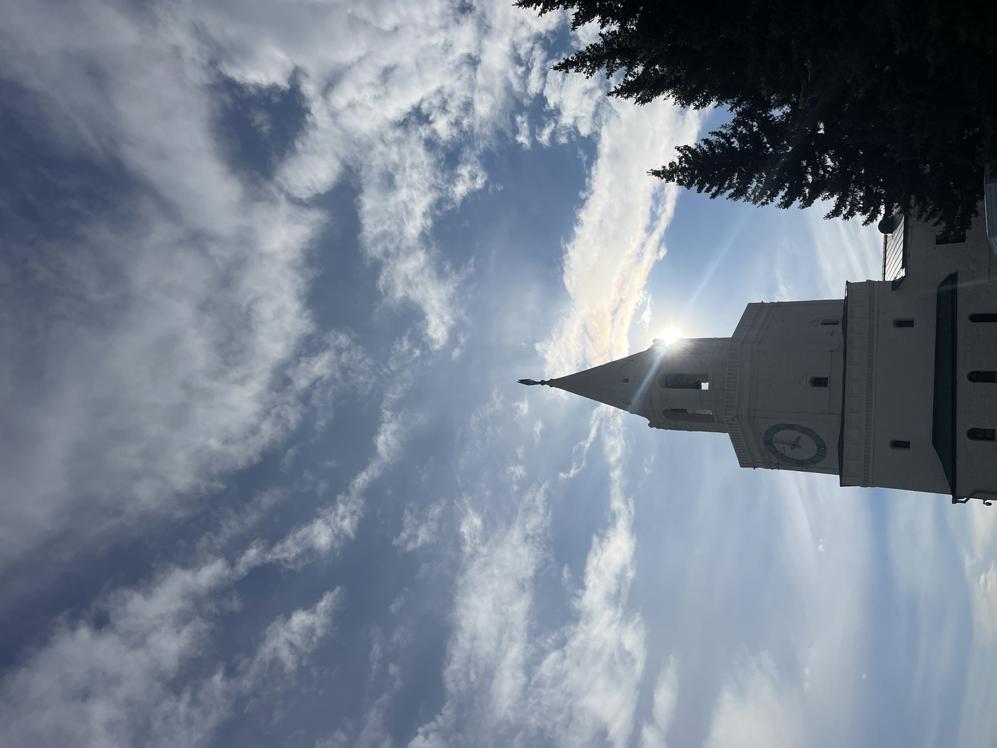 The image displays a low-angle view of a prominent, light-colored architectural tower against a bright blue sky filled with scattered white clouds. The tower, which appears to be a clock tower, features a visible clock face with indistinct Roman numerals. A tapering spire, topped with a finial, extends from the tower's peak. The sun is positioned directly behind the spire and the upper section of the tower, creating a strong sunburst effect and bright lens flare, indicating a sunny daytime scene.
Dark green foliage, likely from evergreen trees, is visible silhouetted against the sky along the top and upper right edges of the frame. The tower's surfaces are well-lit, showcasing its structural details. No people are visible, and no specific activities are taking place. The only visible text is the largely indistinguishable Roman numerals on the clock face. The provided location context for this media is Песчаные Ковали, Russia.