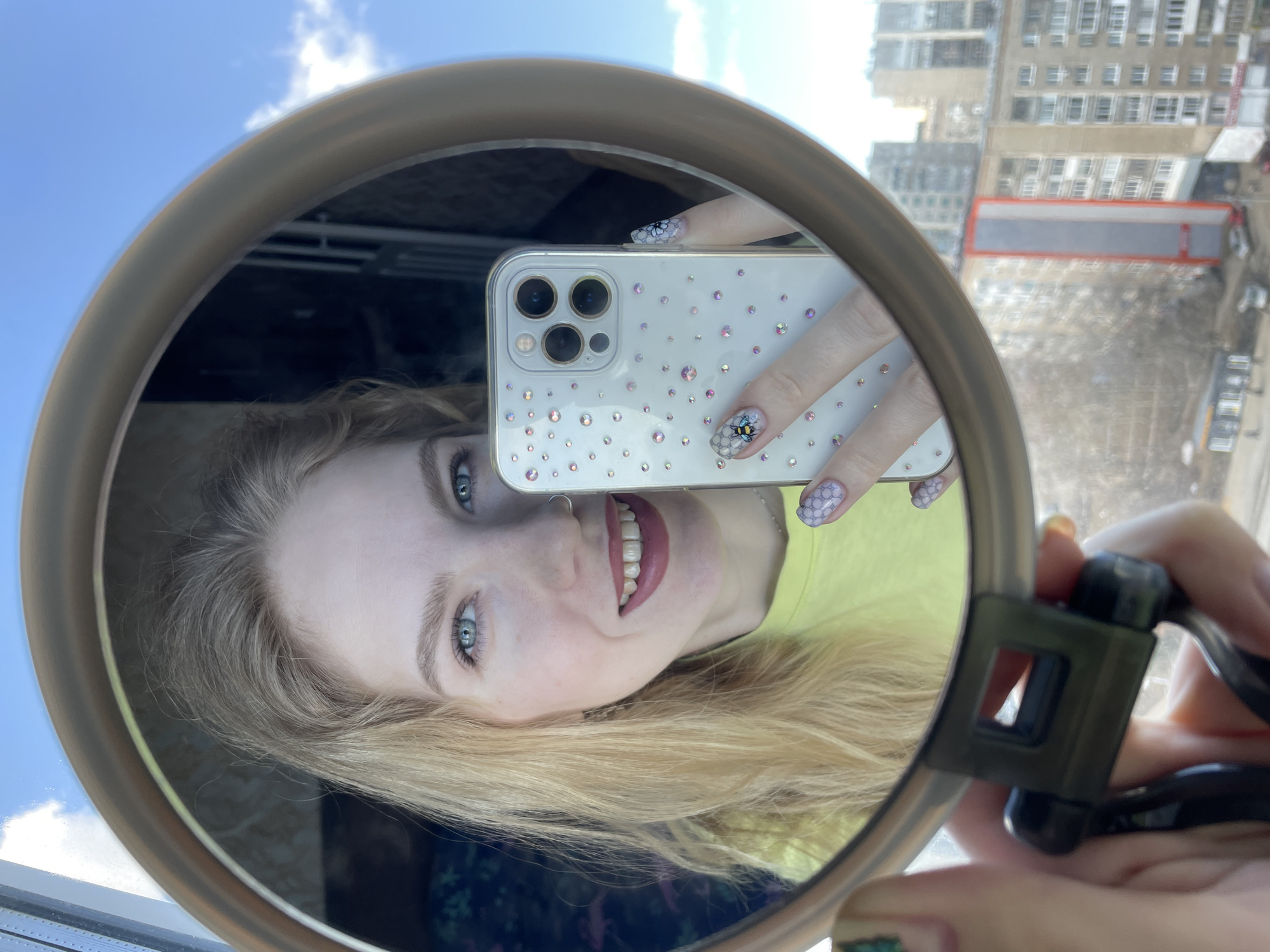 A woman with blond hair and blue eyes is holding a circular mirror, taking a selfie with her phone. The phone case is white with rhinestones. Her nails are decorated with nail art, including one nail with a bee design. The background shows a city skyline with apartment buildings, suggesting a location in an urban area. Given the location mentioned in the prompt, Izhevsk, Russia, this image appears to be taken indoors near a window with a view of the city.