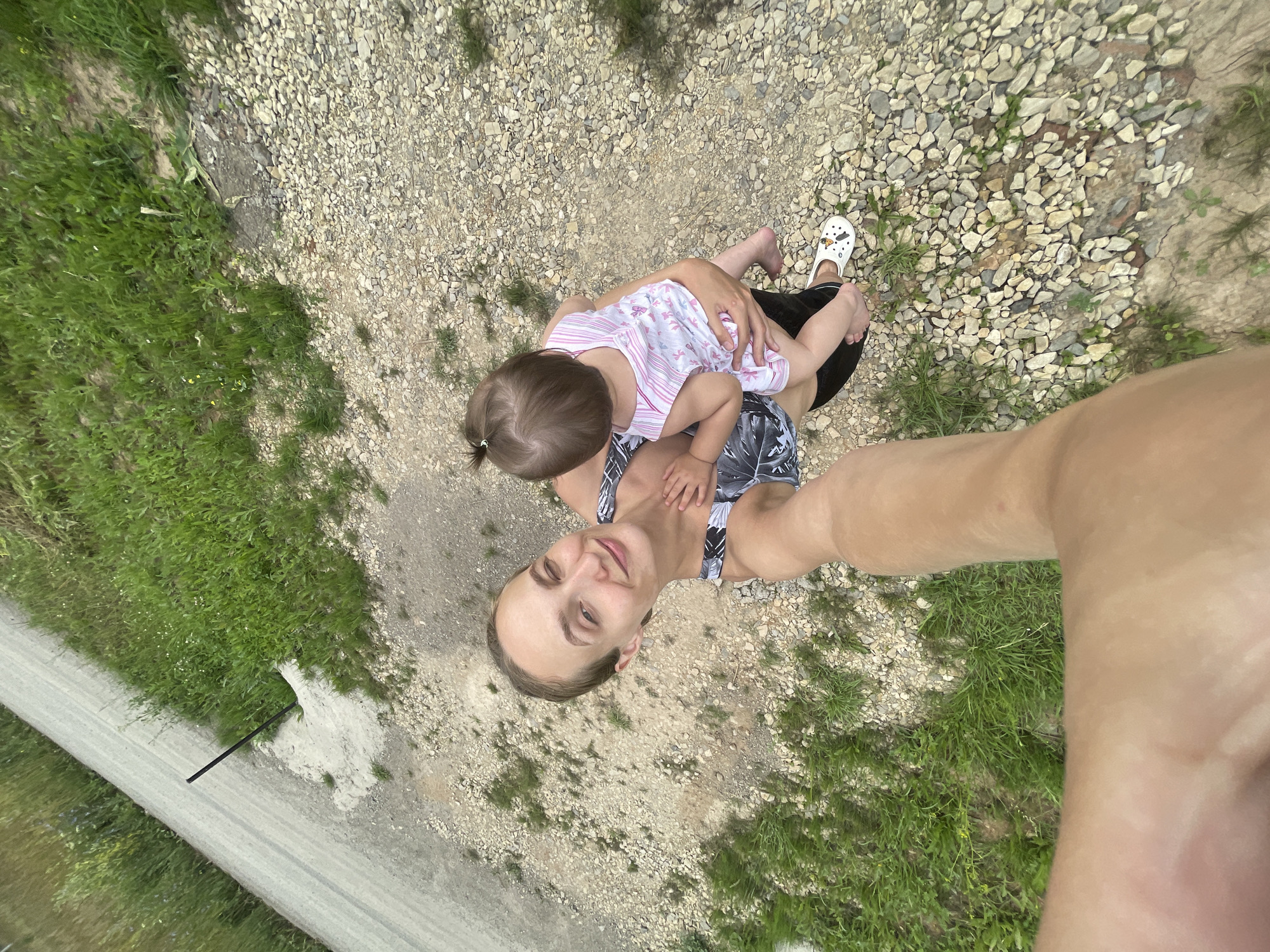 A woman is holding a baby while taking a selfie. The woman is smiling and looking at the camera, and the baby is facing away from the camera. They are standing on a gravel path next to a grassy area, with a dirt road in the background. The scene appears to be outdoors, possibly in a rural setting. The woman is wearing a black and white patterned top and black bottoms, while the baby is wearing a pink and white striped dress. The woman is barefoot, and the baby is wearing white crocs. The location is specified as Podshivalovo, Russia.