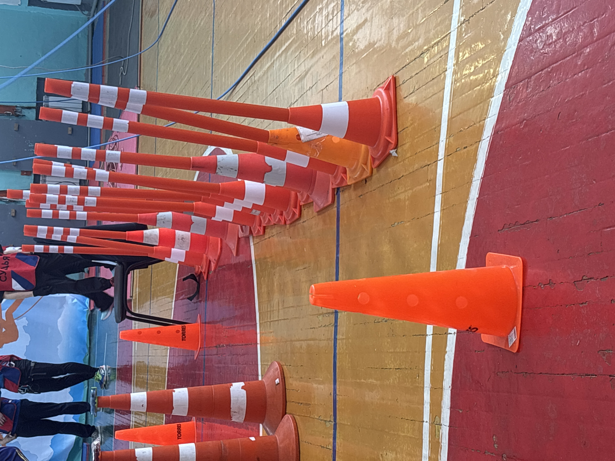 The scene depicts an indoor sports hall in Tayshet, Russia, featuring a wooden floor with distinct red and yellow/orange painted sections, delineated by white and blue lines. In the foreground and midground, a series of approximately twenty orange traffic cones are systematically arranged, some bearing the "TORRES" brand. Interspersed with these are multiple slender orange poles, each marked with white reflective stripes, standing upright from cone bases.
In the background, at least three individuals are partially visible from the waist down. They are dressed in dark trousers and athletic tops, some featuring red, white, and blue patterns, and appear to be wearing harnesses. One individual is positioned on a black folding chair, with the Russian word "СУДЬЯ" (JUDGE) clearly legible on their back. Behind the individuals, a large blue and white banner with graphic designs is displayed against a light blue wall, where blue ropes or cables are also present. The arrangement of cones and poles suggests a structured course or competition.