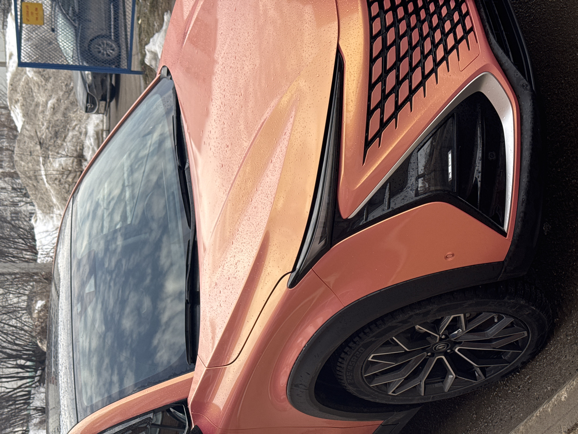 The image depicts the front left quarter of an iridescent orange Omoda C5 crossover SUV in Chaikovsky, Russia. The vehicle's paintwork and windshield are visibly covered in water droplets. It features a modern design with sharp, angular LED daytime running lights integrated into the front fascia, a large black grille with an orange patterned mesh, and prominent black fender flares above dark, multi-spoke alloy wheels. The windshield reflects an overcast sky. The car is parked on a wet, dark paved surface next to a concrete curb. In the background, large piles of dirty snow are visible, indicative of melting conditions. Bare deciduous trees are interspersed behind the snow piles. To the right, a blue chain-link fence separates the foreground from a dark-colored Mazda 6 sedan parked in the distance, with further snow accumulations beyond.