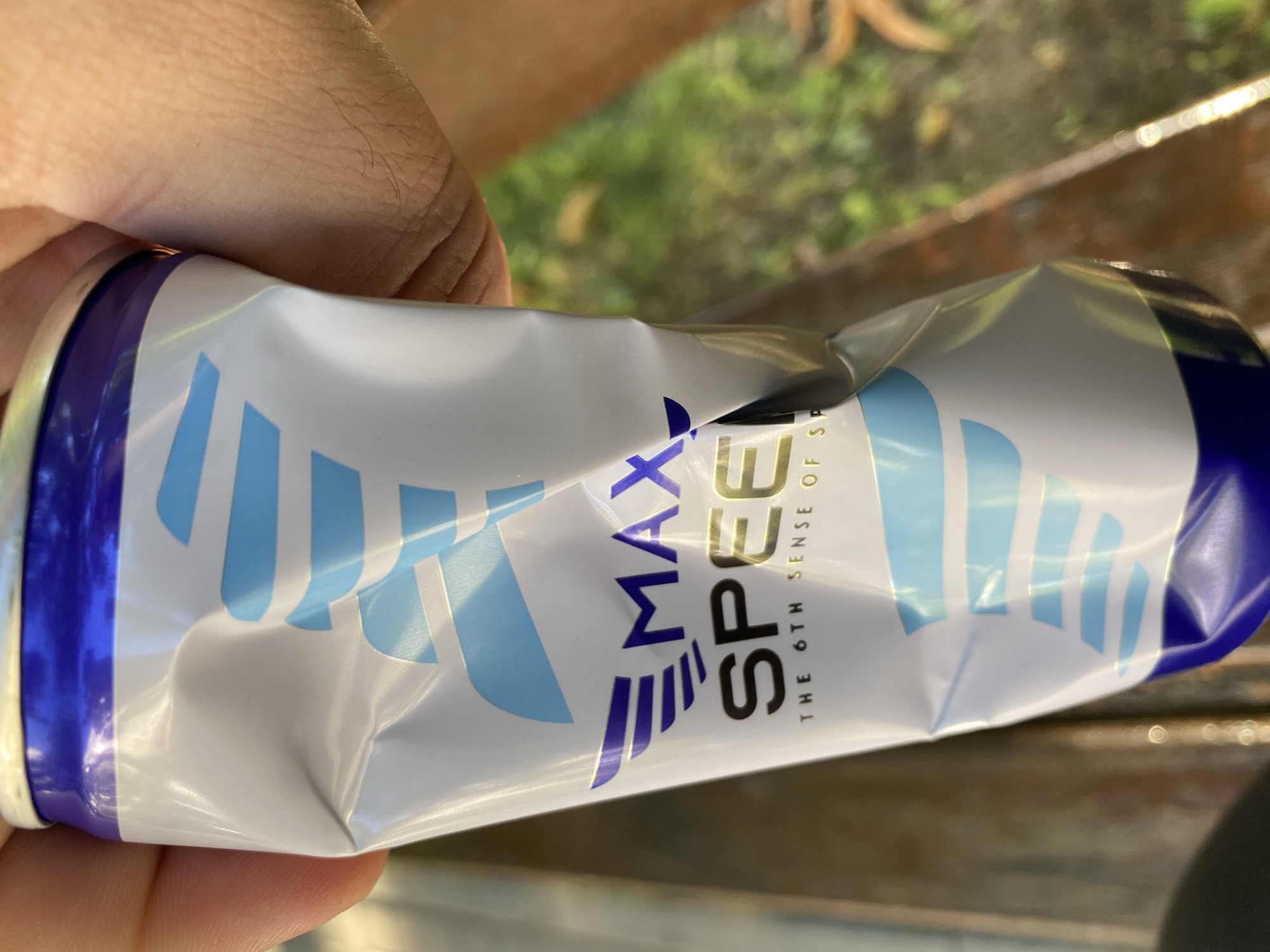 A close-up, low-angle shot shows a crushed aluminum can being held by a person's hand. The can is primarily white with blue and light blue graphic elements, including stylized wings and stripes. The text "MAX SPEED" is prominently displayed in large purple and gold letters, with smaller text below reading "THE 6TH SENSE OF SP". The can is significantly dented and creased, suggesting it has been crushed. The background is softly blurred, showing a mix of green foliage and brown, textured surfaces, possibly suggesting an outdoor setting like a park or garden. The lighting suggests it is daytime. No specific location in Chișinău, Moldova can be identified from the visual information.