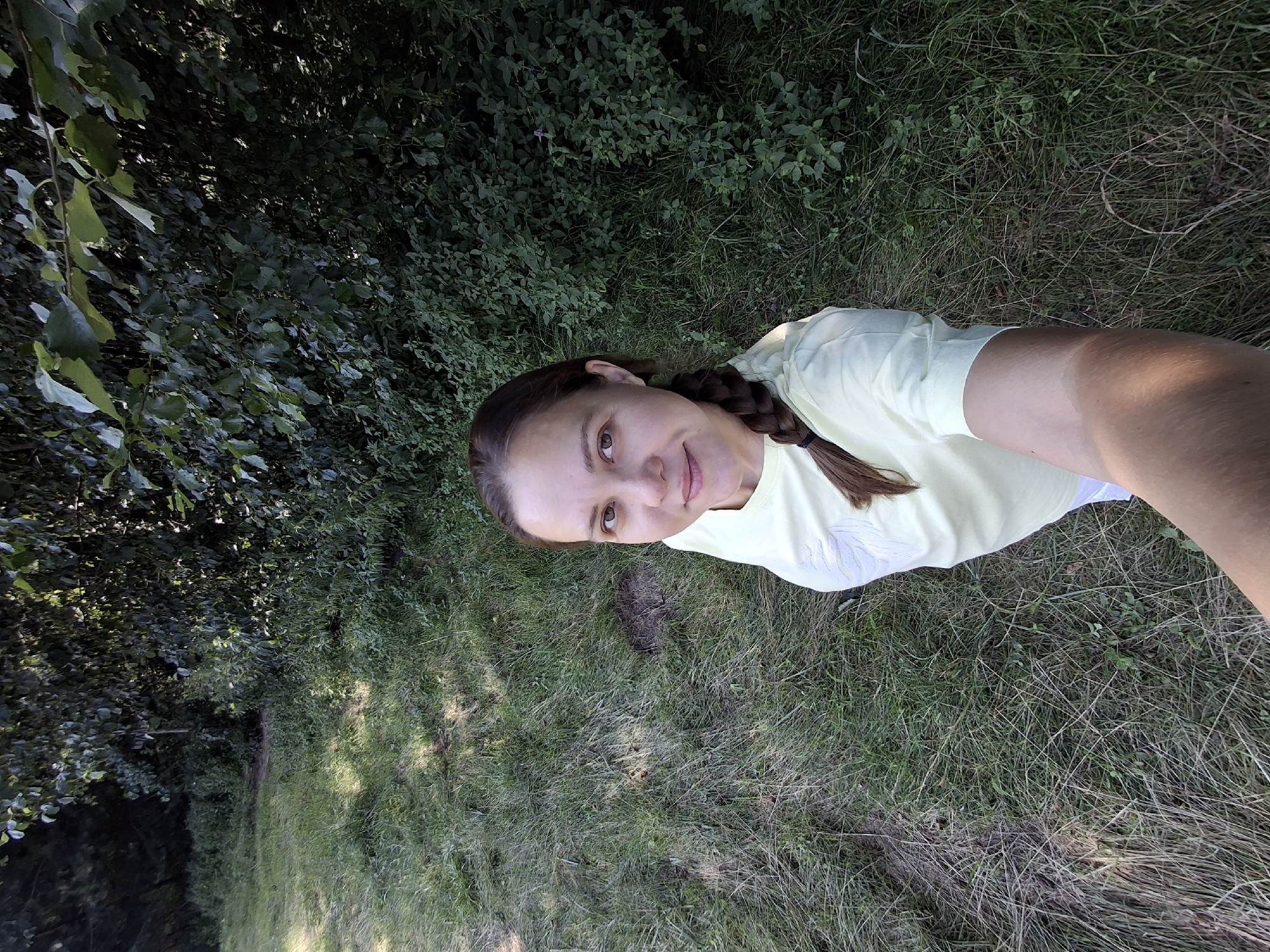 A high-angle, medium shot captures a woman taking a selfie in a grassy, wooded area. The woman, positioned slightly off-center to the right, has her arm extended to capture the photo. She is wearing a light yellow t-shirt and her brown hair is styled in a braid. She looks directly at the camera with a slight smile. The background consists of dense green foliage and trees, with patches of sunlight dappling the grass. The location is in Plishkino, Russia.