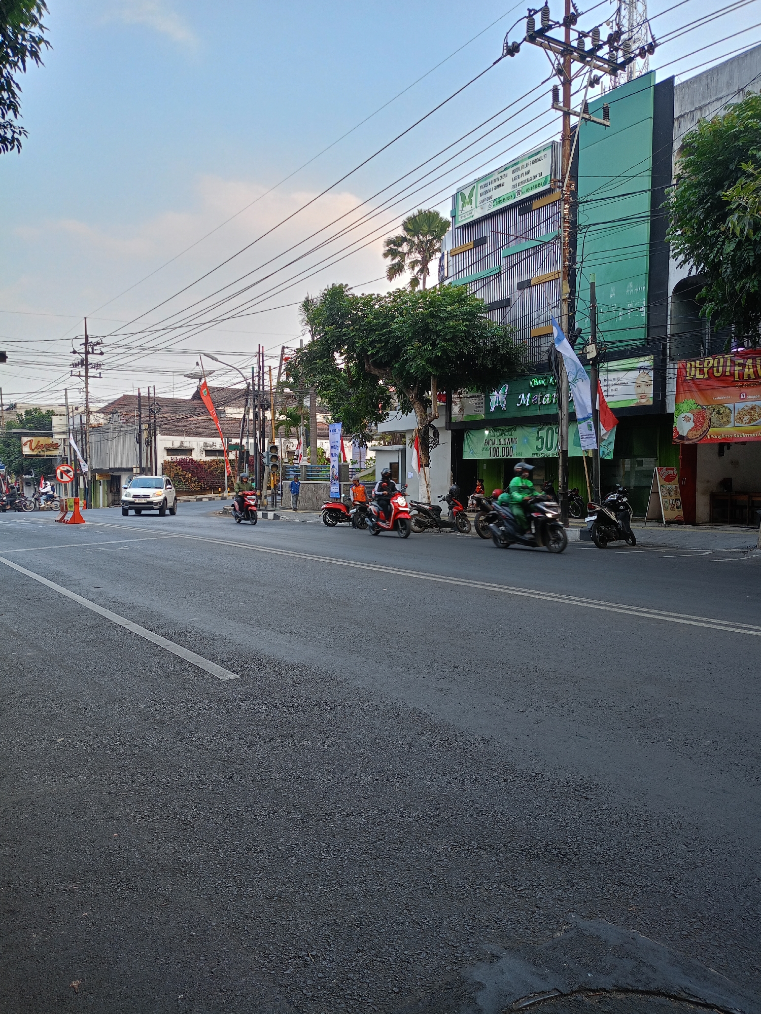 The image captures a street scene in Batu, Indonesia, likely in a commercial area, with a focus on a busy intersection. The foreground shows a paved road with white lane markings. Several motorcycles are in motion, with riders visible on some, suggesting active traffic. In the middle ground, a white car is turning at the intersection, and traffic lights are present. Buildings line the street, including a modern, multi-story green building with signage for "Klinik K" and "Metamorfosis" along with advertisements for facial treatments and discounts. Another building to the right displays a banner for "DEPOT FAVORIT" and appears to be a food establishment. There are also numerous utility poles with a complex network of overhead wires, and flags, some bearing national colors, are visible on poles. Trees and palm trees add to the urban landscape. The sky is partly cloudy with a light blue hue, indicating it is daytime, possibly late afternoon. The overall impression is of a typical urban street in Indonesia with moderate traffic and commercial activity.