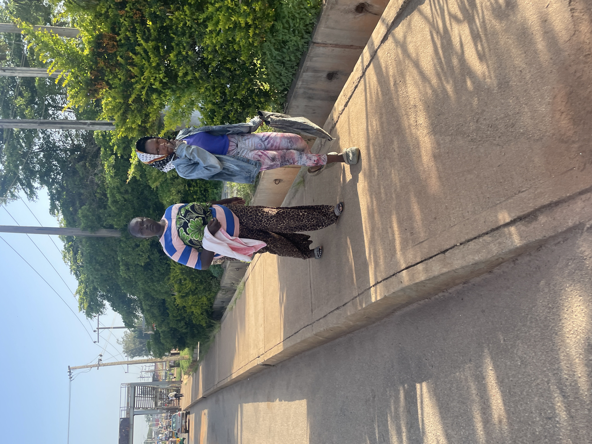 Two women walk side-by-side on a concrete sidewalk adjacent to a paved road in Jos, Nigeria. The woman on the left wears a striped shirt and patterned pants and carries a light-colored bundle. The woman on the right wears a denim jacket, a purple top, and tie-dye patterned leggings, and carries a dark bag. A row of green bushes lines the right side of the sidewalk, and in the distance, a pedestrian bridge and various vehicles are visible on the road. The sky is clear and blue, indicating daylight.