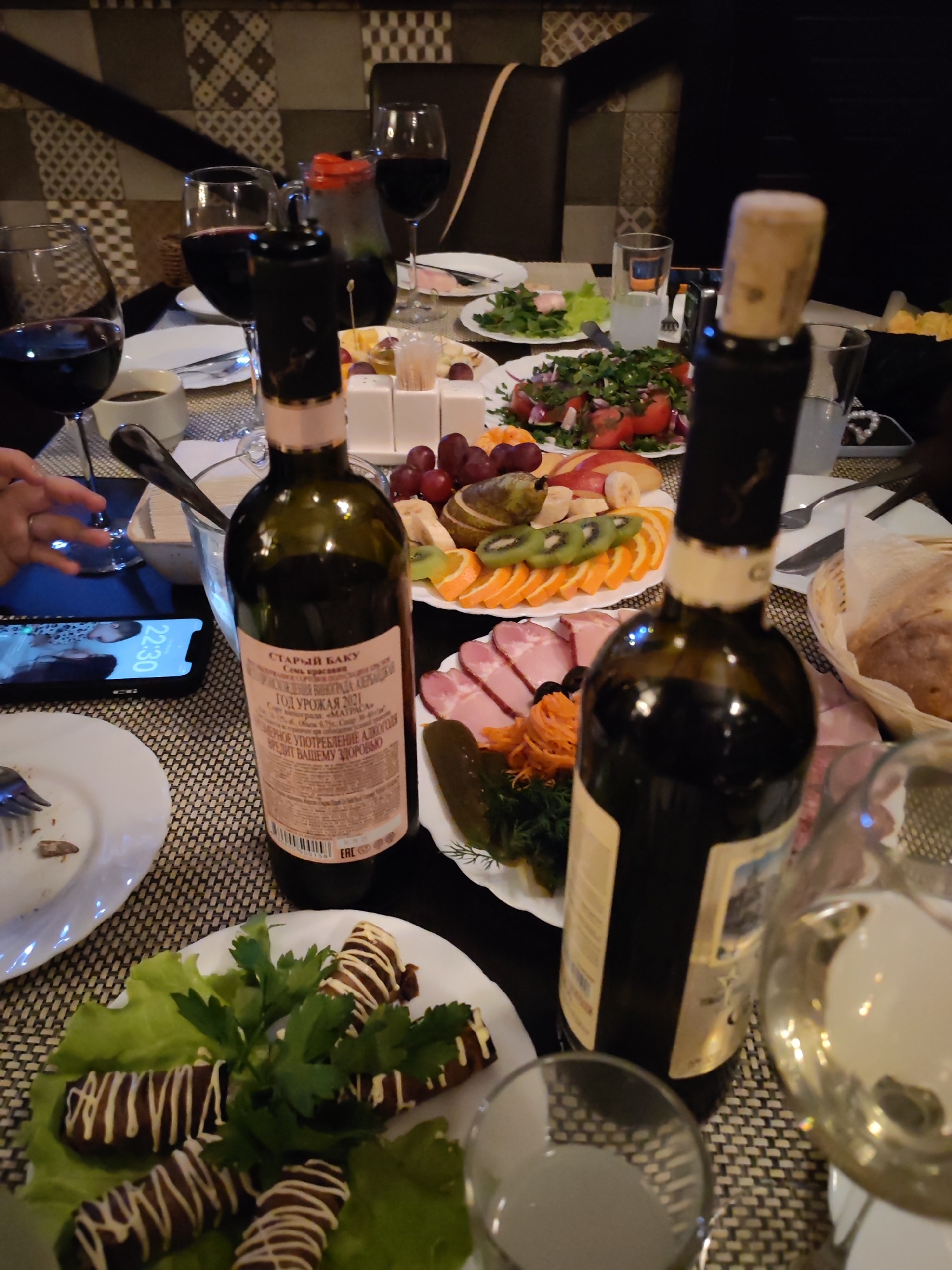 The image captures an indoor dining scene in Izhevsk, Russia, likely during a late evening gathering, indicated by the dim lighting and a smartphone displaying "22:30." The setting features a table laden with various food dishes and drinks, suggesting a social meal or celebration.
In the foreground, two dark glass wine bottles stand prominently. One bottle, closer to the center, has a label mostly visible. Several wine glasses, some filled with red wine, are scattered across the table. A hand is partially visible, reaching for one of the glasses.
The table is set with multiple plates of food. These include a colorful fruit platter with kiwi, oranges, apples, and grapes; a salad with tomatoes and red onions, garnished with fresh herbs; a plate of sliced cured meats and pickles; and another dish featuring dark, elongated food items drizzled with a white sauce, served on lettuce. Bread is visible in a woven basket. The table is covered with a dark, textured placemat or tablecloth. The background shows a dark wall with a patterned design, possibly a decorative panel.
Visible text on the wine bottle label (in Cyrillic) reads:
* "СТАРЫЙ БАКУ" (Old Baku)
* "Семь кранио" (Seven Cranes)
* "ГОД УРОЖАЯ 2021" (Harvest Year 2021)
* "МАТРАСА" (Matrasa)
* "ВЕРНОЕ УПОТРЕБЛЕНИЕ АЛКОГОЛЮ ВРЕДИТ ВАШЕМУ ЗДОРОВЬЮ" (Correct consumption of alcohol harms your health – a standard health warning).