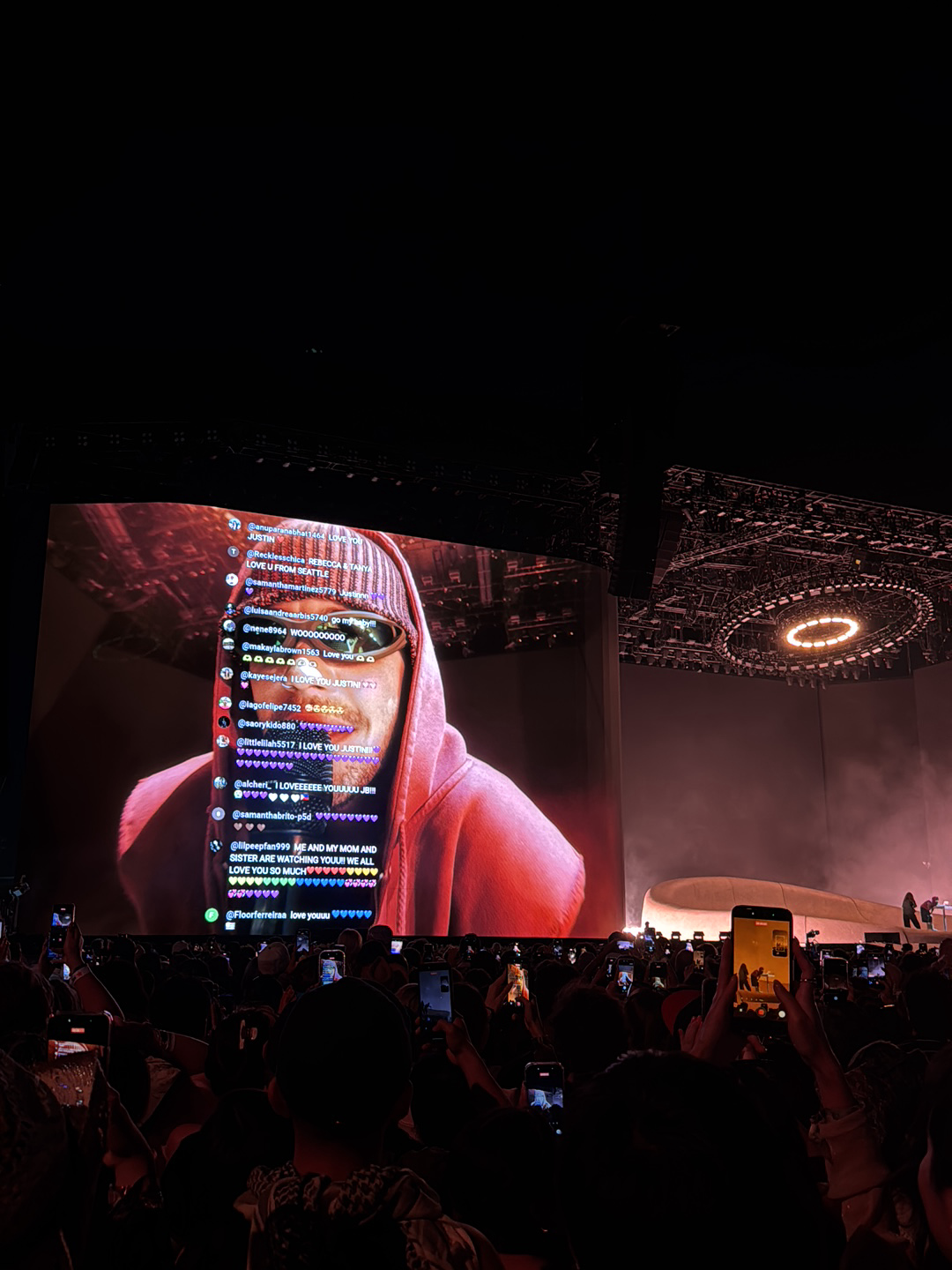 The image shows a concert at an event in Indio, United States, with a large crowd holding up phones to capture the performance. The focal point is a giant screen displaying a close-up of a person wearing sunglasses and a red hoodie. The screen also shows live comments from viewers. Overhead, there is a complex structure of lights and rigging. The foreground consists of the audience, visible as a sea of dark figures holding up their phones.