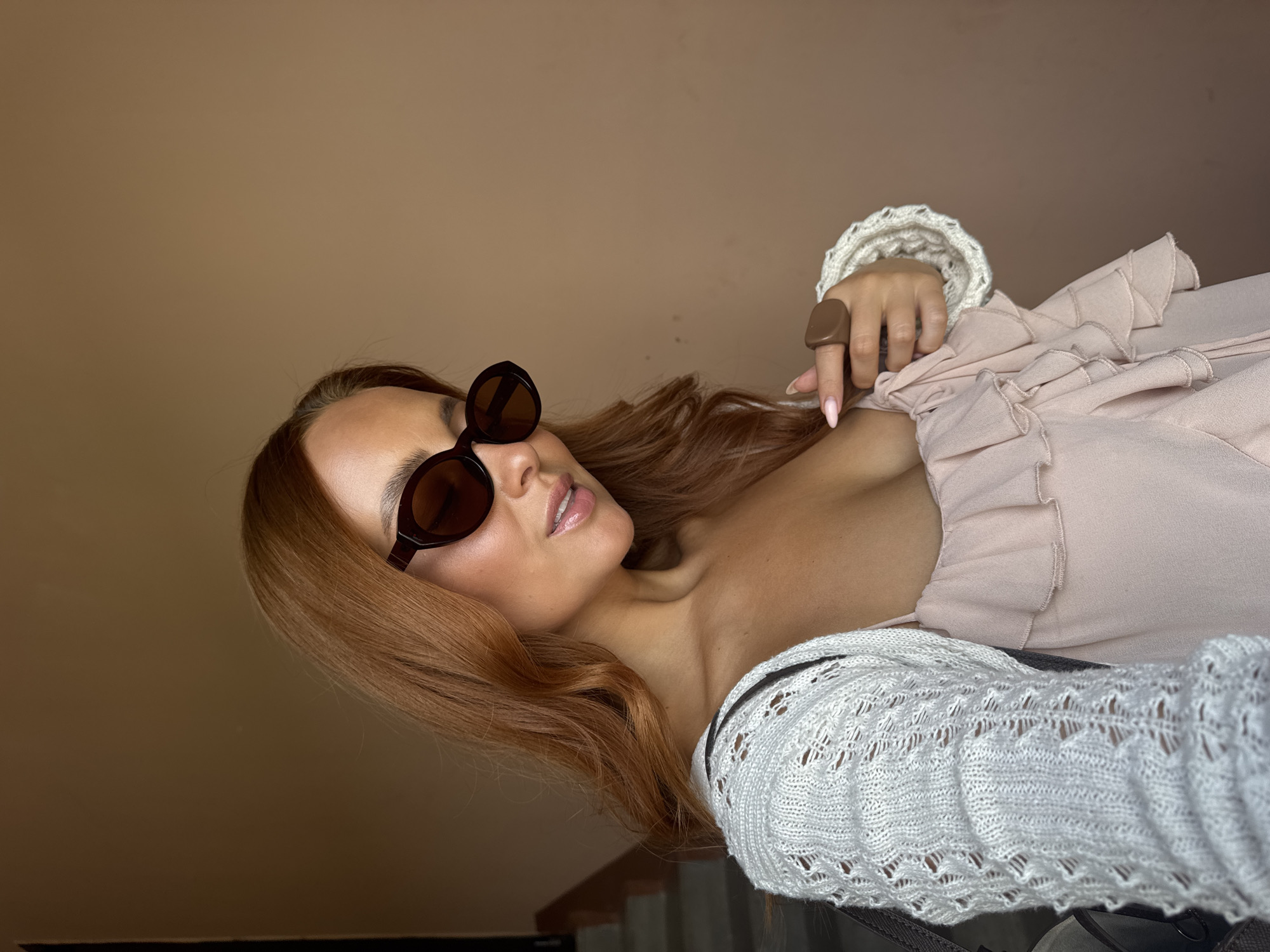 A woman with auburn hair and sunglasses is seen from the chest up, leaning her head down and to the right. She is wearing a pale pink, ruffled camisole and a white crocheted cardigan. A large, brown ring is on her right hand, which is resting on her chest. The background is a plain, light brown wall. There are no elements in the image that suggest a location in Chaikovsky, Russia.