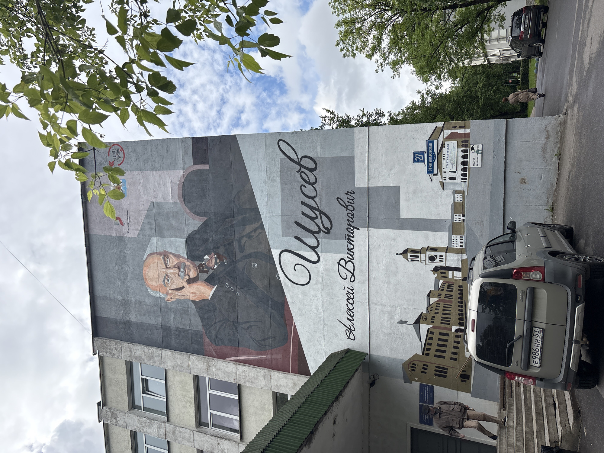Here's a detailed description of the image:
**Scene:** The image shows a building facade in Veliky Novgorod, Russia, adorned with a large mural featuring a portrait of Alexei Viktorovich Shchusev. Shchusev, an architect, is depicted in a seated pose, holding his face with his right hand. Alongside his portrait, the mural includes a cityscape featuring historical buildings and the address "Stratilatovskaya Street, 27."
**Environment:** The scene is set outdoors on a partially sunny day. A branch with green leaves is visible in the upper left corner of the image, suggesting a tree nearby. The sky is partly cloudy. A street is visible to the side of the building, including a grey car.
**Objects:** Key objects in the image include:
* The mural itself, featuring Shchusev's portrait and cityscape elements.
* The building facade, including windows and a green-colored overhang above an entrance.
* A grey-colored car parked beside the building. The license plate number is E986 HH 53.
**People:**
* A person is walking down stairs near the building entrance. They appear to be wearing a military style jacket and pants.