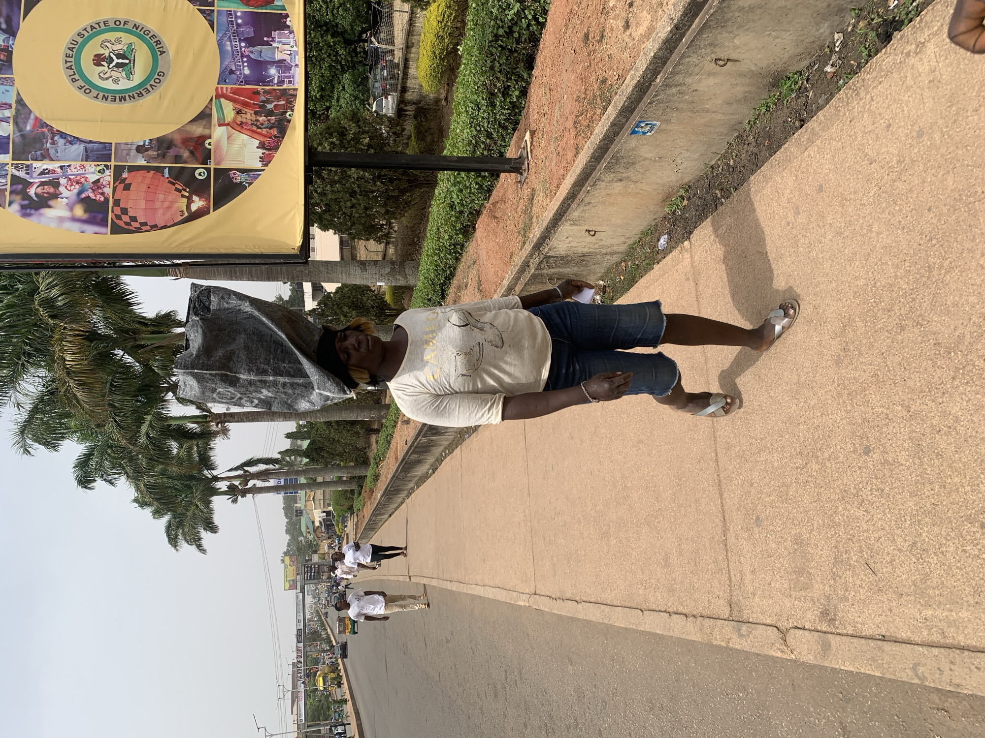 The image, captured in Jos, Nigeria, depicts a daytime street scene with a focus on a person standing on a sidewalk. The individual, with dark skin, is wearing a light-colored t-shirt with a graphic, blue denim shorts, and silver-toned sandals. A dark, possibly patterned, fabric is draped over their head and left shoulder. They are looking towards the camera, holding a small white object in their right hand.
To the immediate left of the person are several tall palm trees, and beyond them, the hazy sky is visible. To their right stands a large, yellow billboard-like structure. This billboard prominently features a circular emblem with the text "PLATEAU STATE OF NIGERIA" and "GOVERNMENT OF PLATEAU STATE," along with the Nigerian coat of arms. Around this central emblem, various smaller images are displayed, including a hot air balloon, people at what looks like a performance, and other local scenes.
The paved sidewalk stretches into the distance, running parallel to a wide road. Further down the road, several other people are visible, some of whom appear to be walking in groups, with a few individuals dressed in matching white shirts. Vehicles are also faintly discernible in the background. The street features a concrete curb and a median strip with sparse vegetation. Bright daylight illuminates the scene, casting shadows on the ground. A small blue sign is attached to the concrete barrier along the sidewalk, but its text is illegible. In the far distance, a banner with the partially visible text "THE SONS OF GLORY" can be seen on the right side of the road. The overall impression is that of a bustling street in an urban environment.