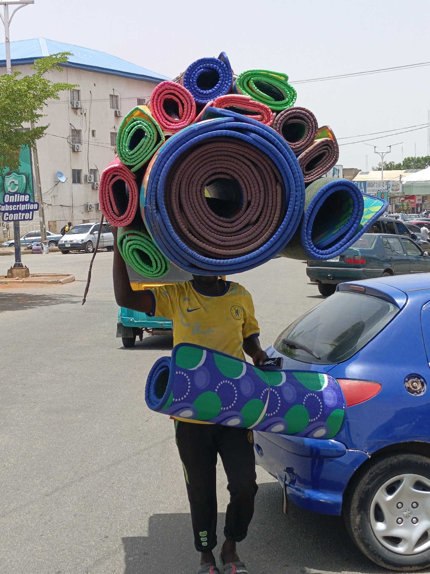 A man in a yellow Chelsea T-shirt is carrying a stack of rolled-up mats on his head and another mat under his arm in a city. The mats are colorful and vary in size. He stands next to a blue car parked on a paved street. There are other cars and buildings in the background, and a sign that says "Online Subscription Control". The location is Unknown, Unknown.