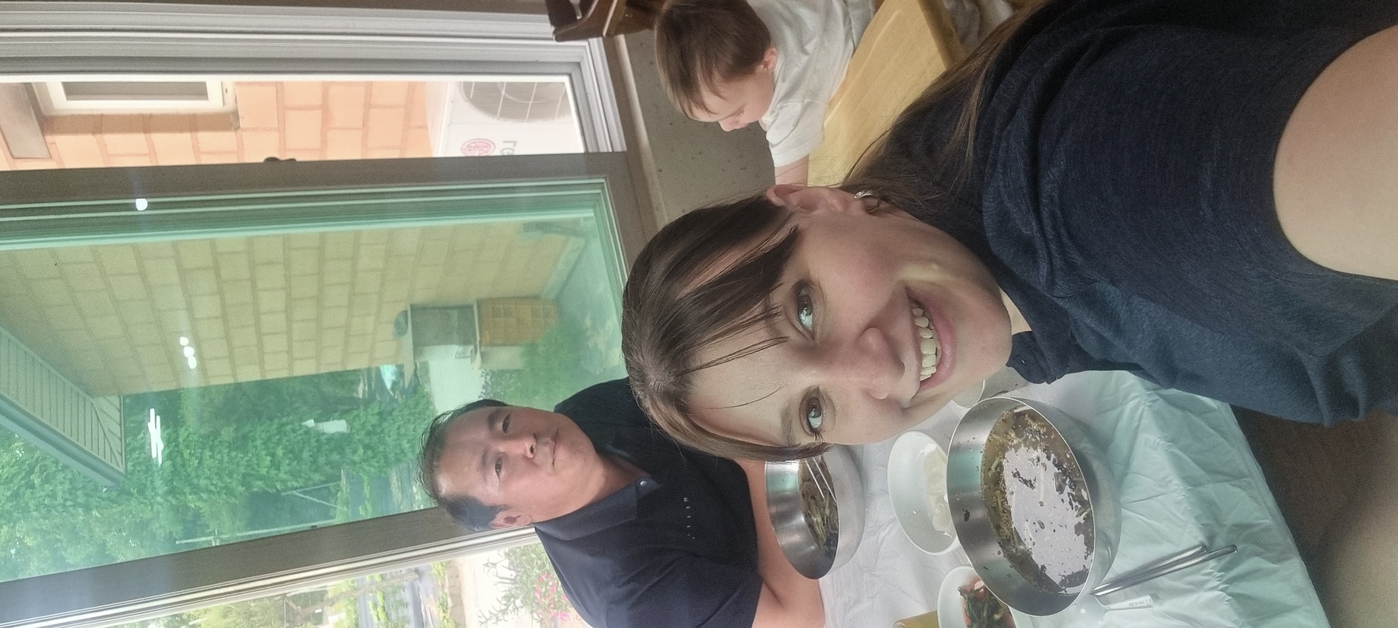 An informal indoor photo taken in Dunnae-myeon, South Korea, captures three individuals around a dining table. The image, oriented vertically, features a smiling woman in the foreground with dark hair and bangs, wearing a dark blue t-shirt, looking directly at the camera. To her left, a man with short dark hair, dressed in a dark polo shirt, also looks towards the camera with a neutral expression. A child with light brown hair and a white t-shirt is partially visible in the background, looking downwards.
The table in front of them is covered with a white tablecloth over a light blue-green sheet and holds several Korean-style metal bowls, some filled with a liquid dish containing green elements. Smaller white bowls and metal chopsticks are also arranged on the table, indicating a meal in progress.
Large windows behind the table offer a view of the outdoors. Through the glass, an LG-branded outdoor air conditioning unit is distinctly visible, along with brick-textured building exteriors and abundant green foliage. Reflections of indoor lighting and a patterned ceiling or awning can also be seen in the windowpanes. The presence of natural light suggests the scene takes place during the daytime. The overall setting is a casual dining environment.