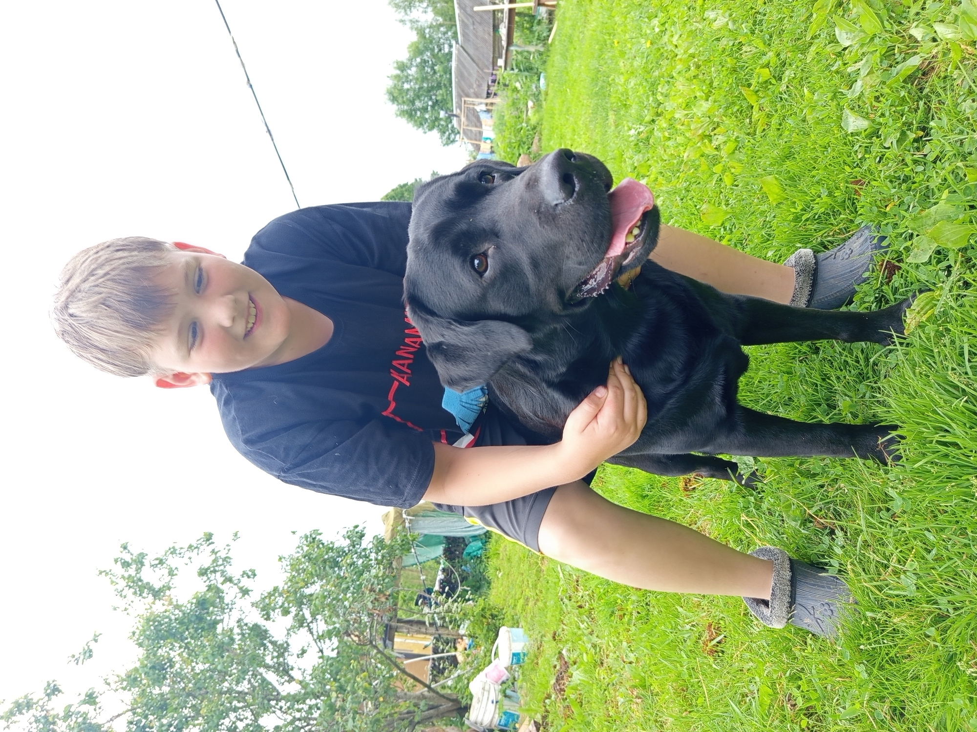 The image shows a young boy and a black Labrador dog sitting on lush green grass outdoors. The boy is wearing a black t-shirt and dark shorts, and the dog is sitting in his lap. The dog's tongue is out, and it appears to be panting happily. The boy is smiling and looking at the camera, with his arm around the dog.
In the background, there are some trees and a rustic wooden structure, possibly a shed or a small house, suggesting a rural or backyard setting. The sky is overcast, indicating a cloudy day. The overall mood is one of joy and companionship between the boy and his dog.
There is some visible text on the boy's t-shirt, which appears to be in red letters, partially obscured by the dog. The text seems to include the letters "ANAN". The image captures a warm moment between a child and his pet on a summer day. The location is Ostrovtsy, Russia.