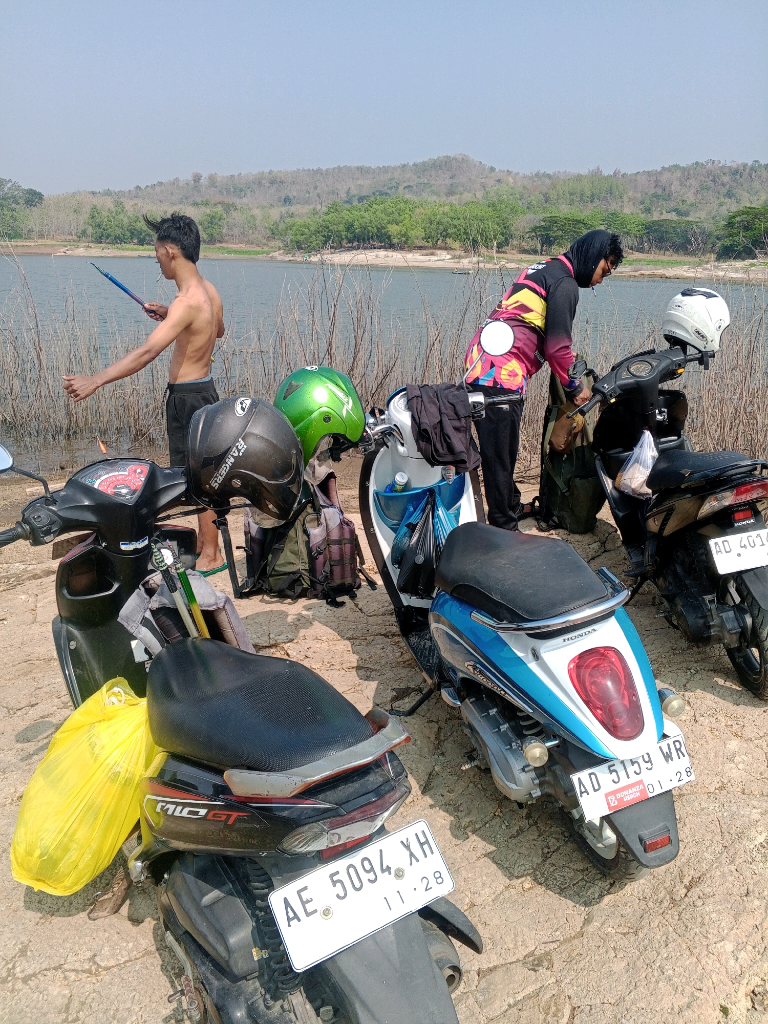 In Wonogiri, Indonesia, on a sunny day, two individuals are present near a body of water. A young man, shirtless and wearing dark shorts, stands in shallow water and holds a fishing rod. He is also smoking a cigarette. Beside him, a person wearing a hooded jacket with a colorful pattern leans on a scooter. Several scooters are parked on a dirt path near the water's edge. Helmets are placed on the scooters, and bags are visible, including a bright yellow bag and backpacks. The landscape behind them features dry vegetation and a distant, tree-covered hill under a clear sky.
