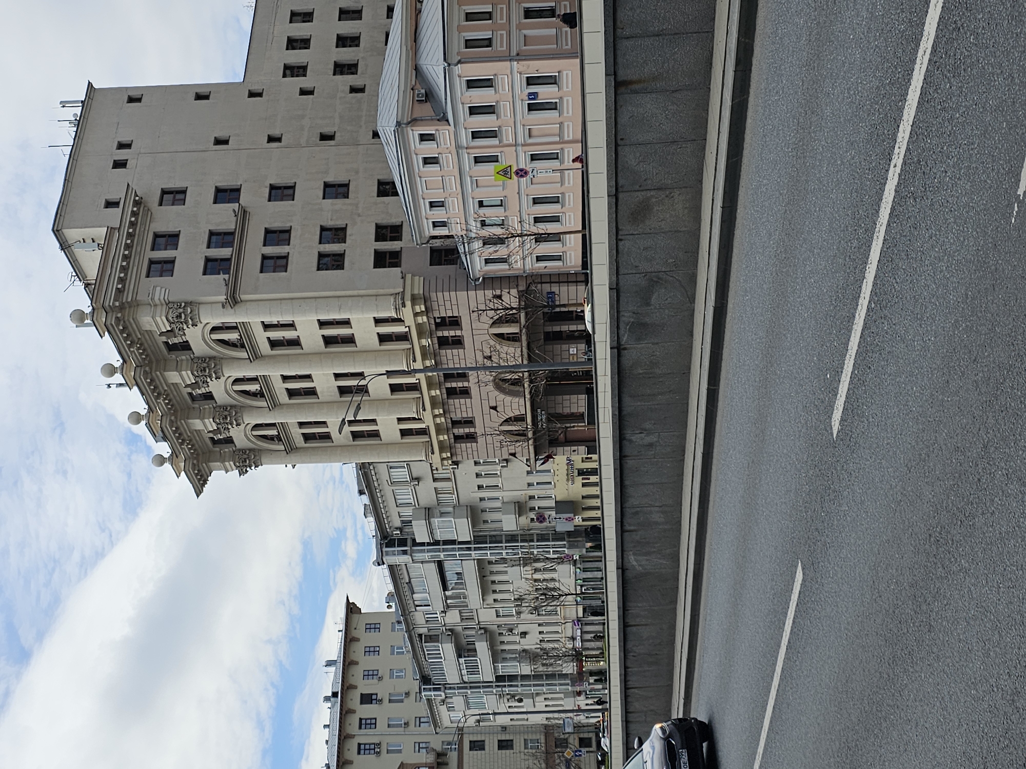 The image shows a street in Moscow, Russia, with buildings on one side and a roadway on the other. The buildings are a mix of architectural styles, including Soviet-era and more modern designs. A multi-lane road is visible, marked with painted lines. A car can be seen on the road. The sky is partly cloudy.