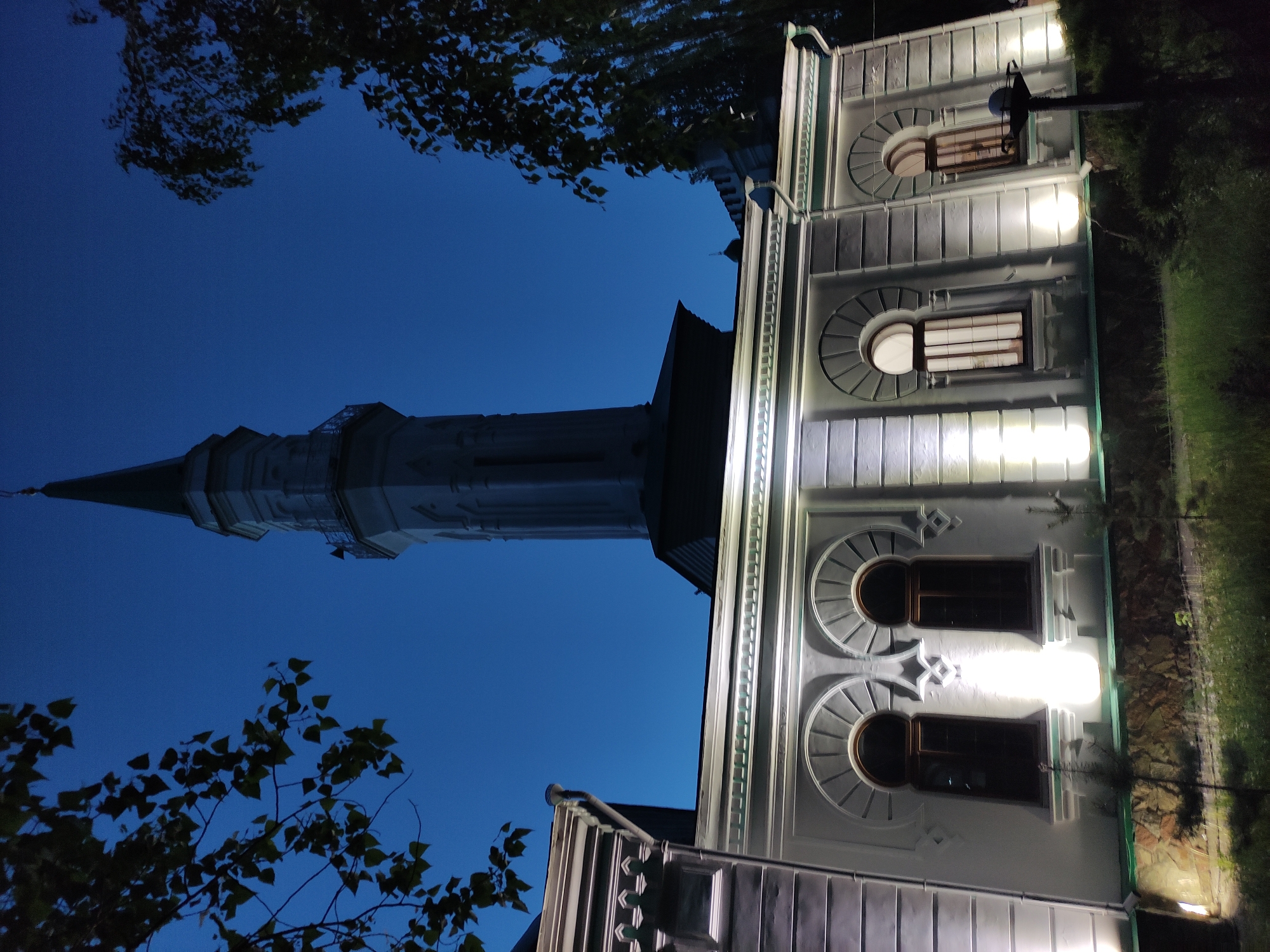The image depicts the Masjid named after Mashkhur Zhusup in Pavlodar, Kazakhstan, at night. The minaret is illuminated and stands tall against the dark blue sky. The building's facade is lit, showcasing architectural details like arched windows and decorative elements. Tree branches are visible in the foreground.