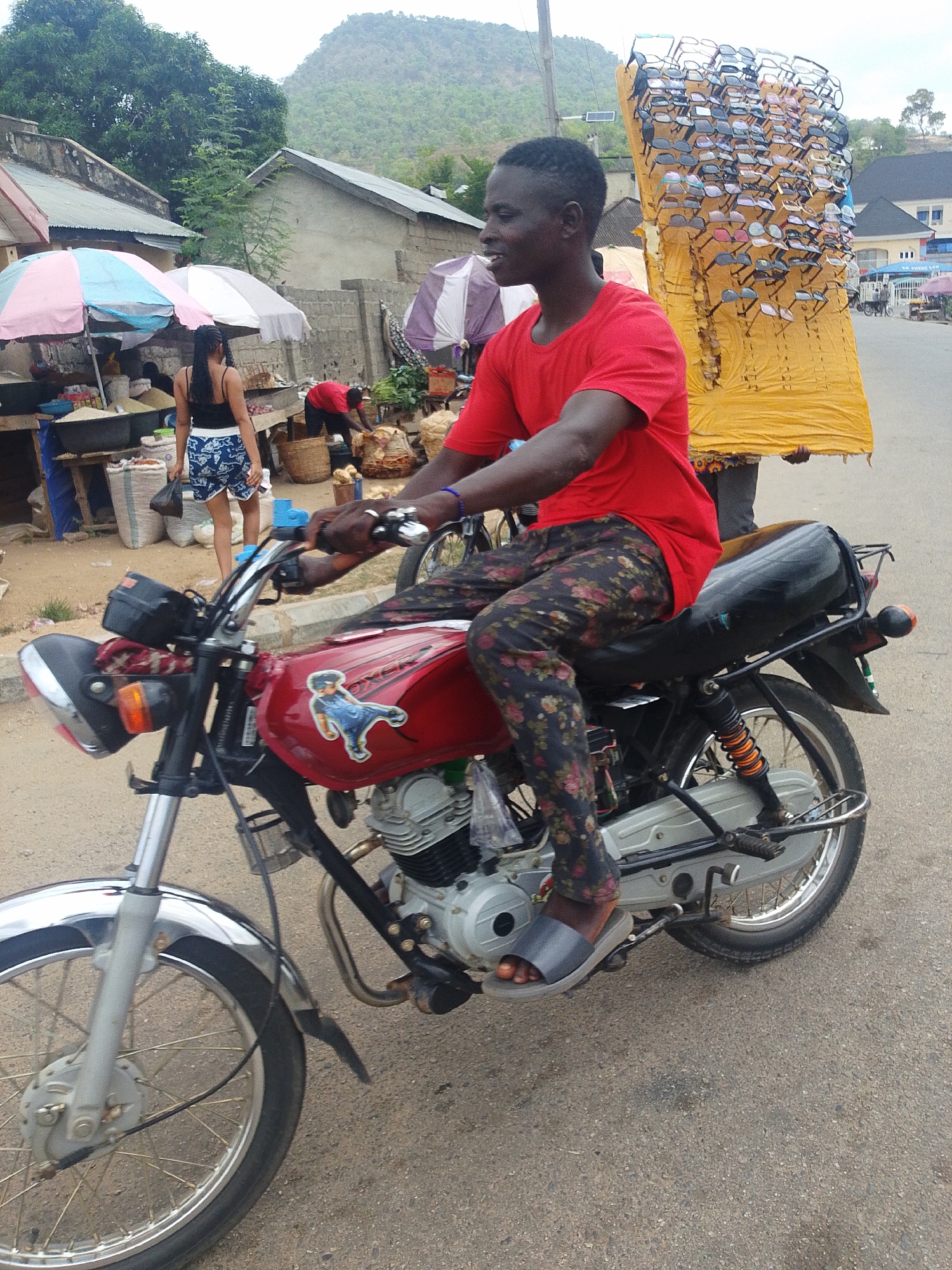 The image shows a man riding a red motorcycle on a street in a town, possibly in Unknown, Unknown. The man is wearing a red t-shirt, floral patterned pants, and sandals. Behind him, another person carries a large yellow sheet covered with eyeglasses. In the background, there are buildings, market stalls with umbrellas, and a woman in shorts and a tank top walking away from the market. A mountain is visible in the distance.