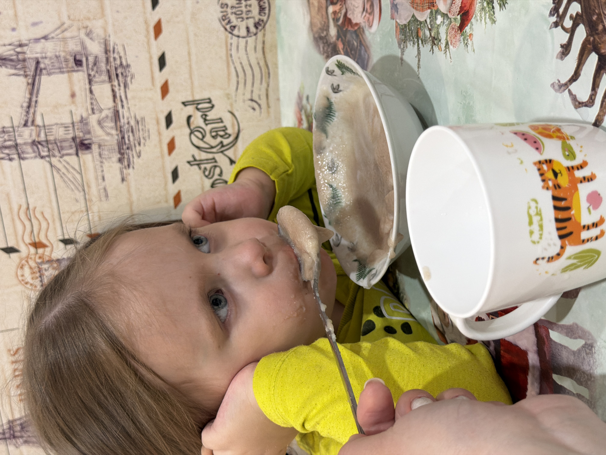 A child is being fed from a spoon. The child has light brown hair and blue eyes, and is wearing a bright yellow shirt with a black pattern. The child is lying on a surface with a pattern resembling old postcards and stamps. A white bowl with a light-colored, possibly oatmeal-like substance sits next to the child. A white mug with animal and fruit designs is also present. The image was taken indoors. No further information indicates the location as Chaikovsky, Russia.