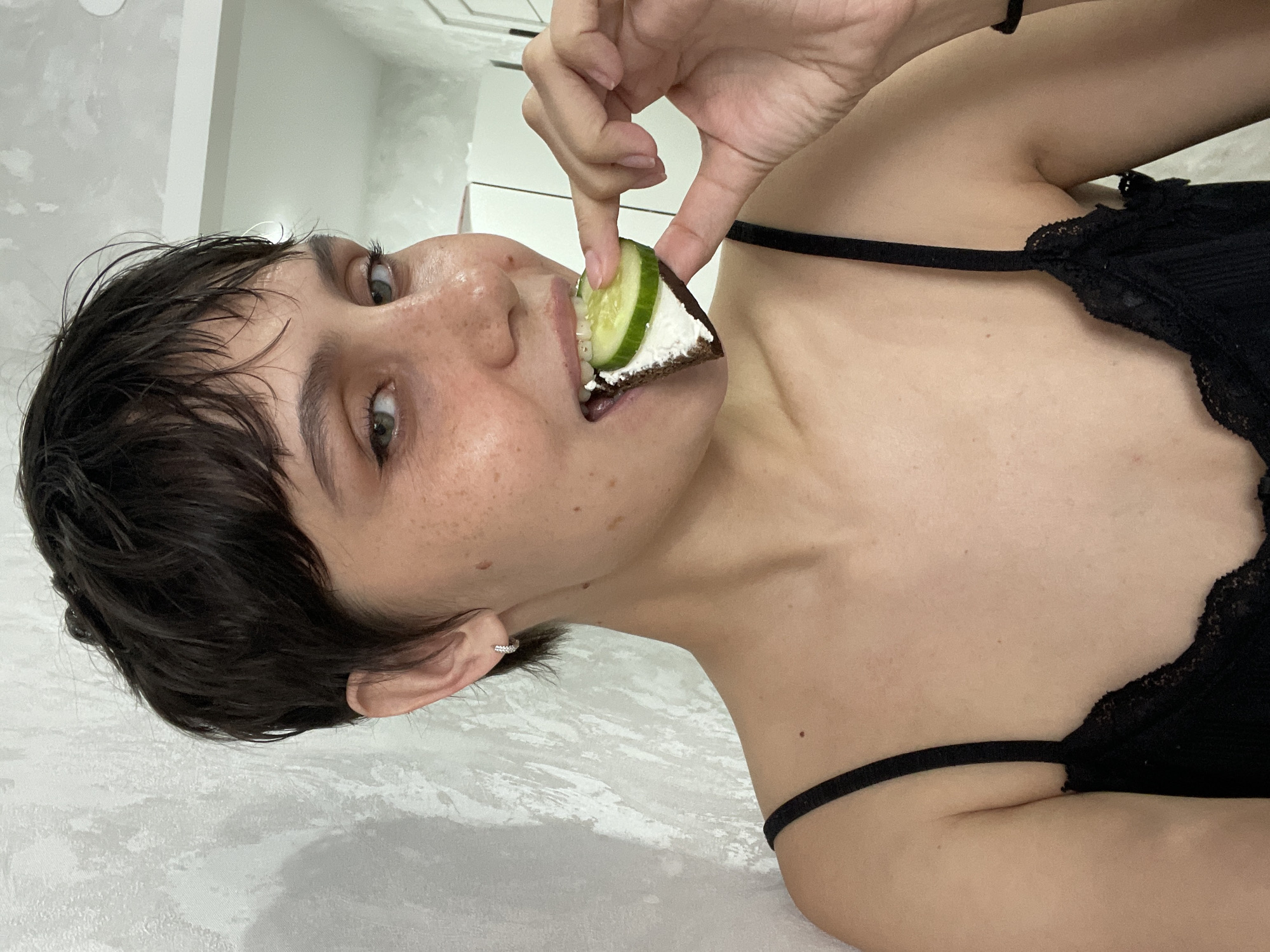 A woman with short dark hair is holding a small piece of bread topped with white cheese and a slice of cucumber to her mouth. She is wearing a black tank top. The background is a light grey surface with a slight texture.